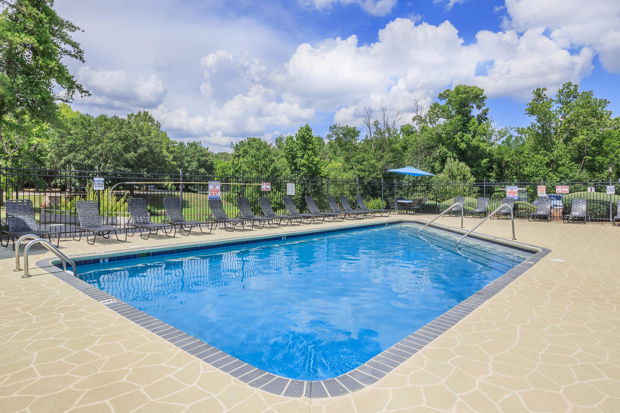 A refreshing outdoor swimming pool surrounded by a tan stone deck. Poolside lounge chairs are neatly arranged along the perimeter, and a vibrant blue sky with fluffy white clouds is visible above. Lush green trees line the background, creating a serene atmosphere.