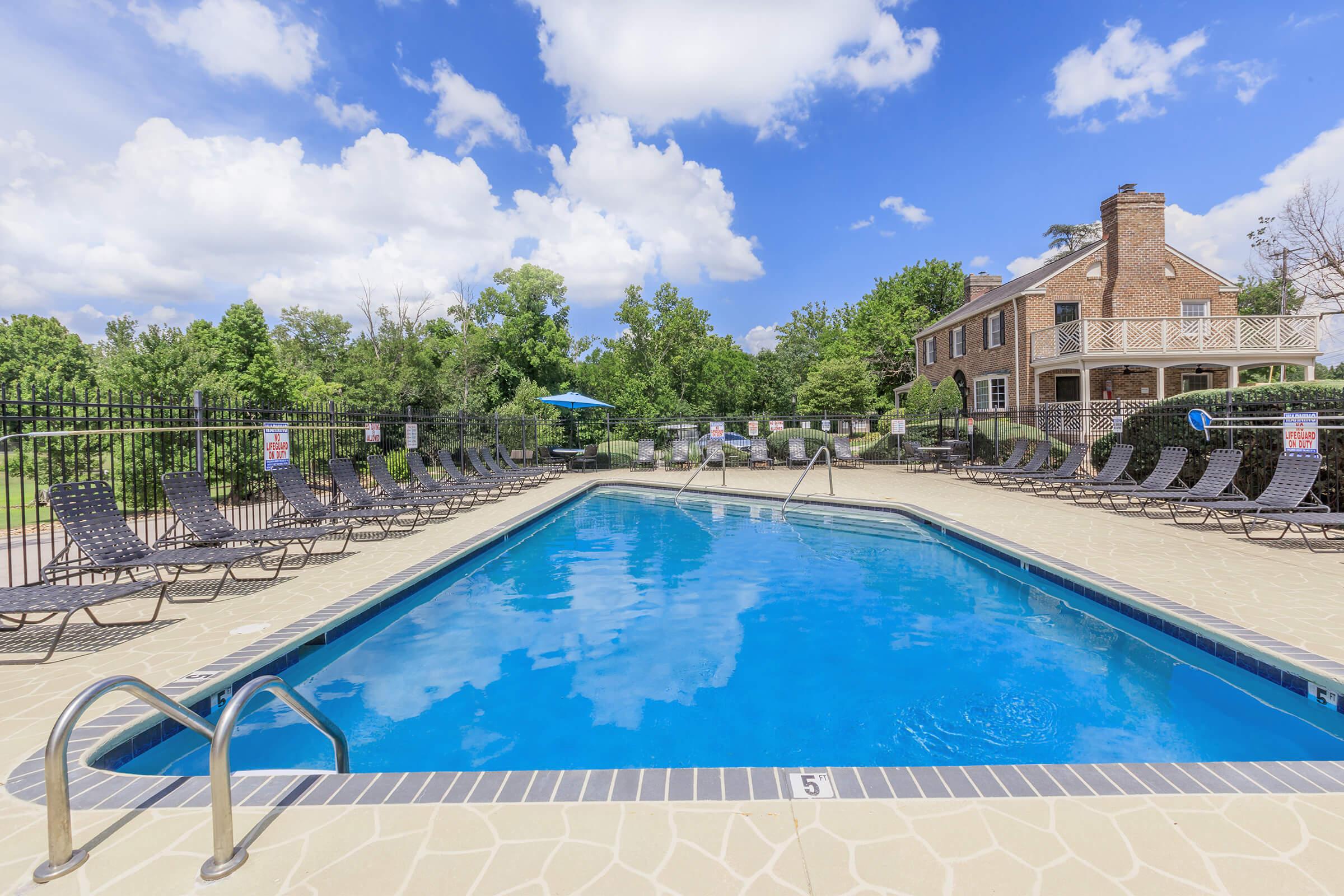 A clear blue swimming pool surrounded by lounge chairs, with a backdrop of green trees and a large brick building. The sky is bright with fluffy white clouds, and a sun umbrella is visible near the pool area, creating a relaxing outdoor setting.