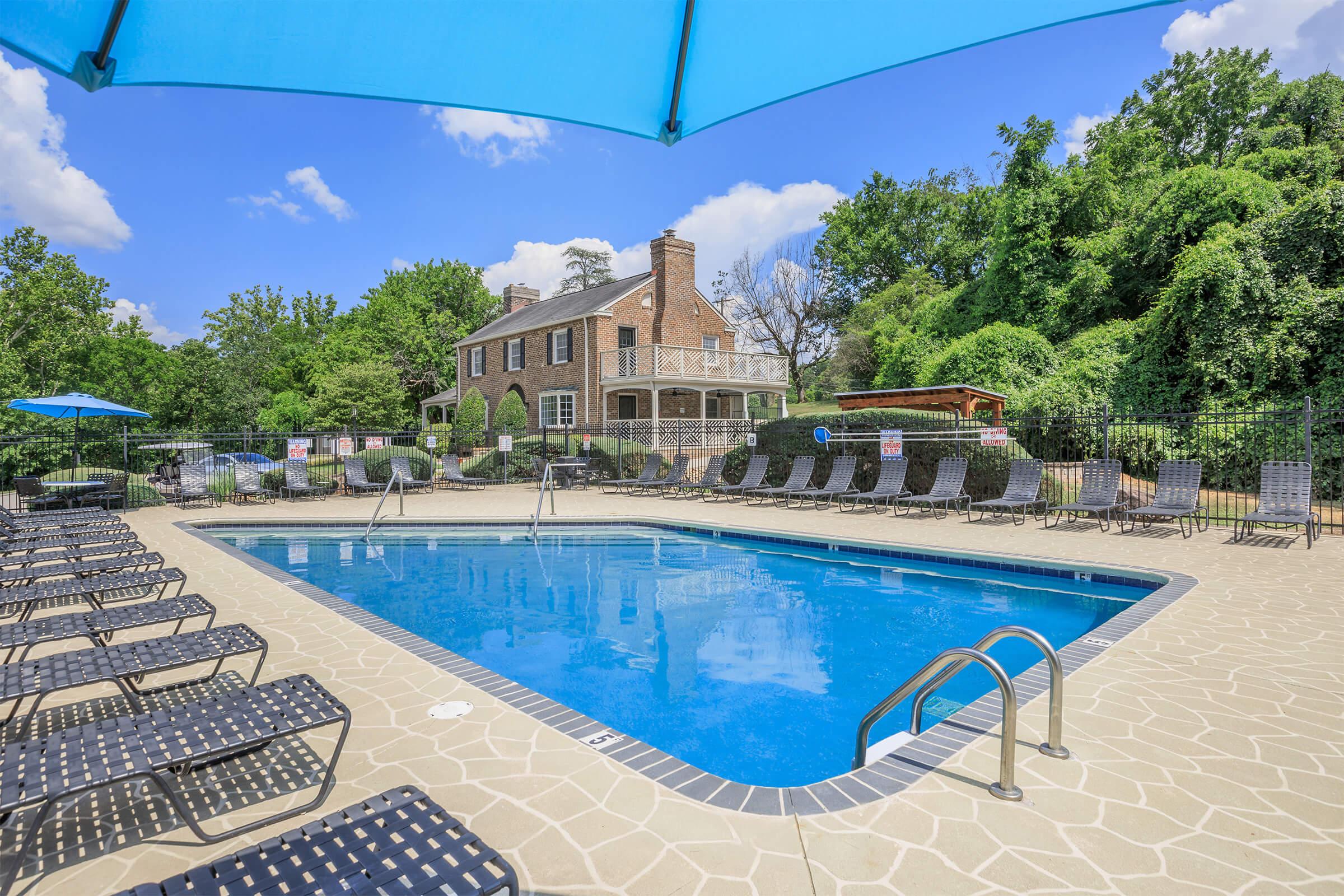 A swimming pool with a landscaped area in the background featuring a large brick house. Lounge chairs line the poolside, and a large blue umbrella provides shade. Lush greenery surrounds the area, with a clear blue sky and fluffy clouds overhead, creating a serene outdoor setting.