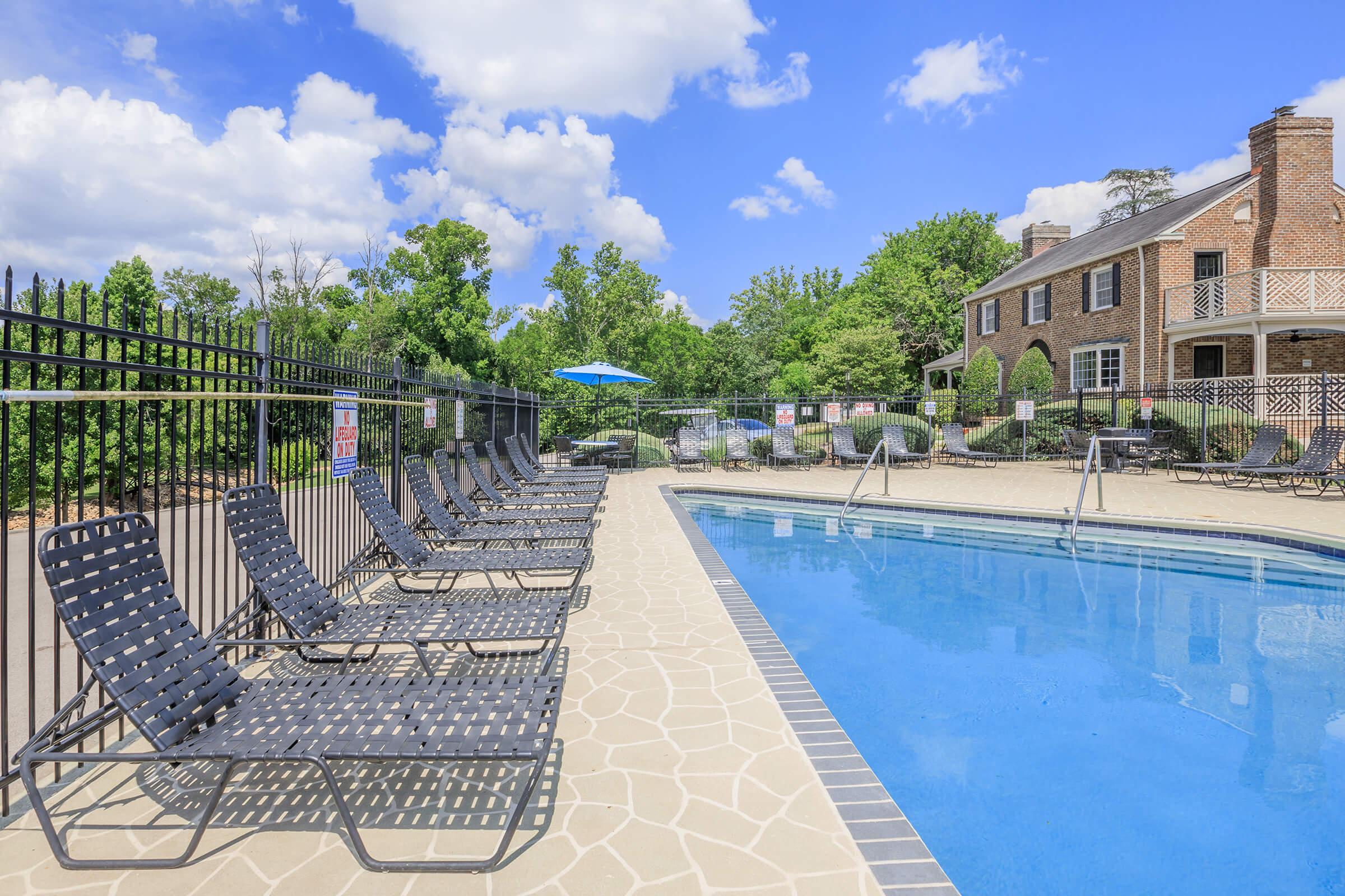 A clear, sunny day at a poolside area featuring a blue swimming pool surrounded by lounge chairs. In the background, there's a brick building with a patio and trees lining the perimeter. A blue umbrella adds a touch of color. The atmosphere is inviting and perfect for relaxation.