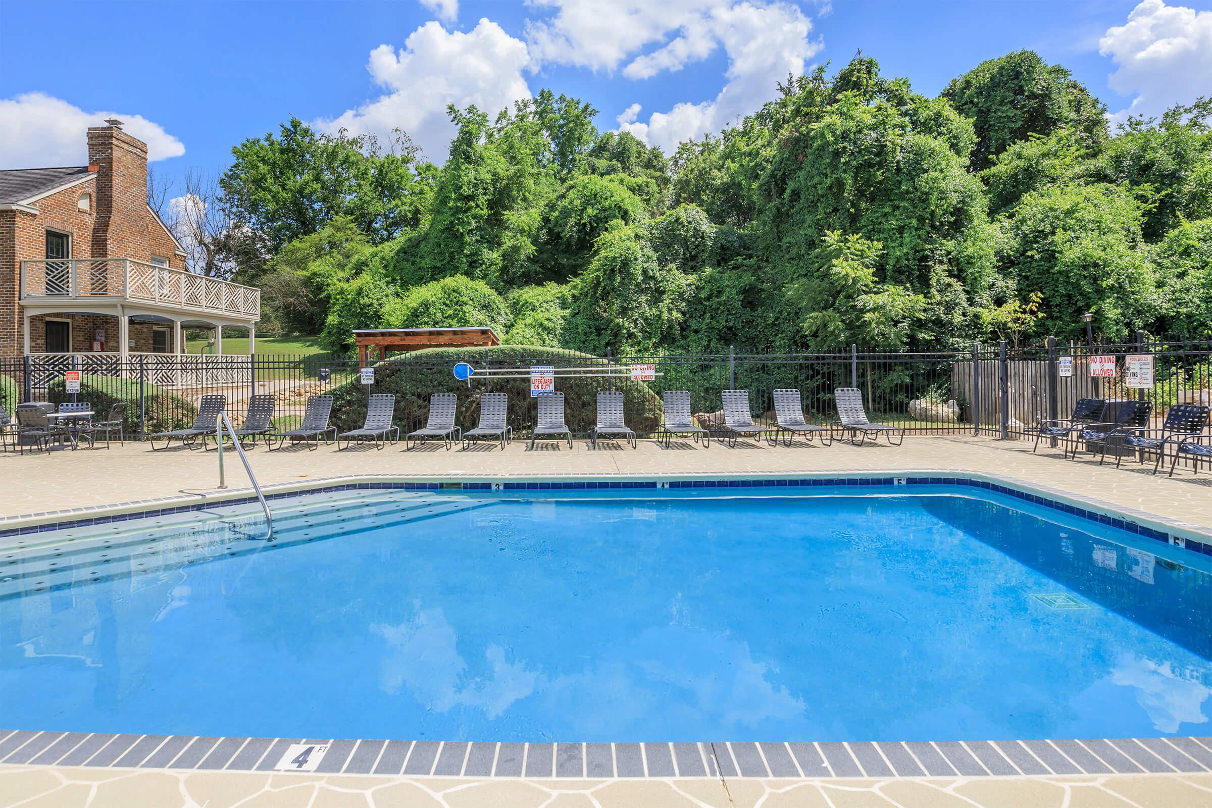 A clear blue swimming pool surrounded by lounge chairs, with lush green trees and bushes in the background. A fence encloses the pool area, and there are signs posted around the perimeter. The sky is bright with fluffy white clouds.