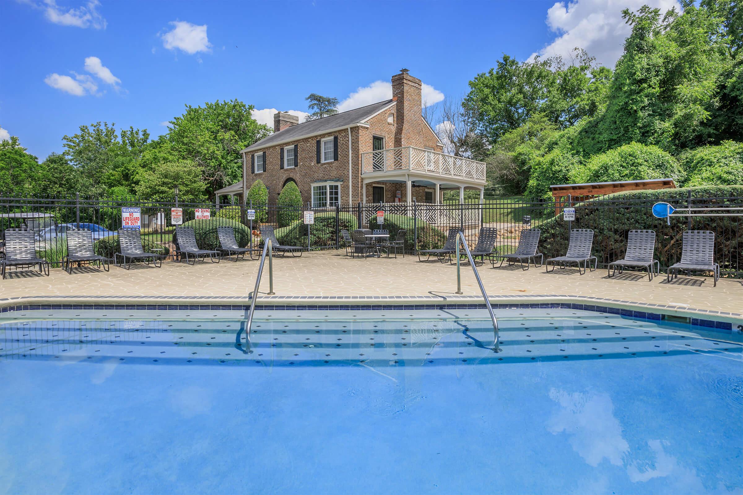 A clear blue swimming pool in the foreground with steps leading in, surrounded by lounge chairs. In the background, there’s a two-story brick house with a balcony, set among lush greenery and a blue sky with scattered clouds. The area is enclosed by a black fence.
