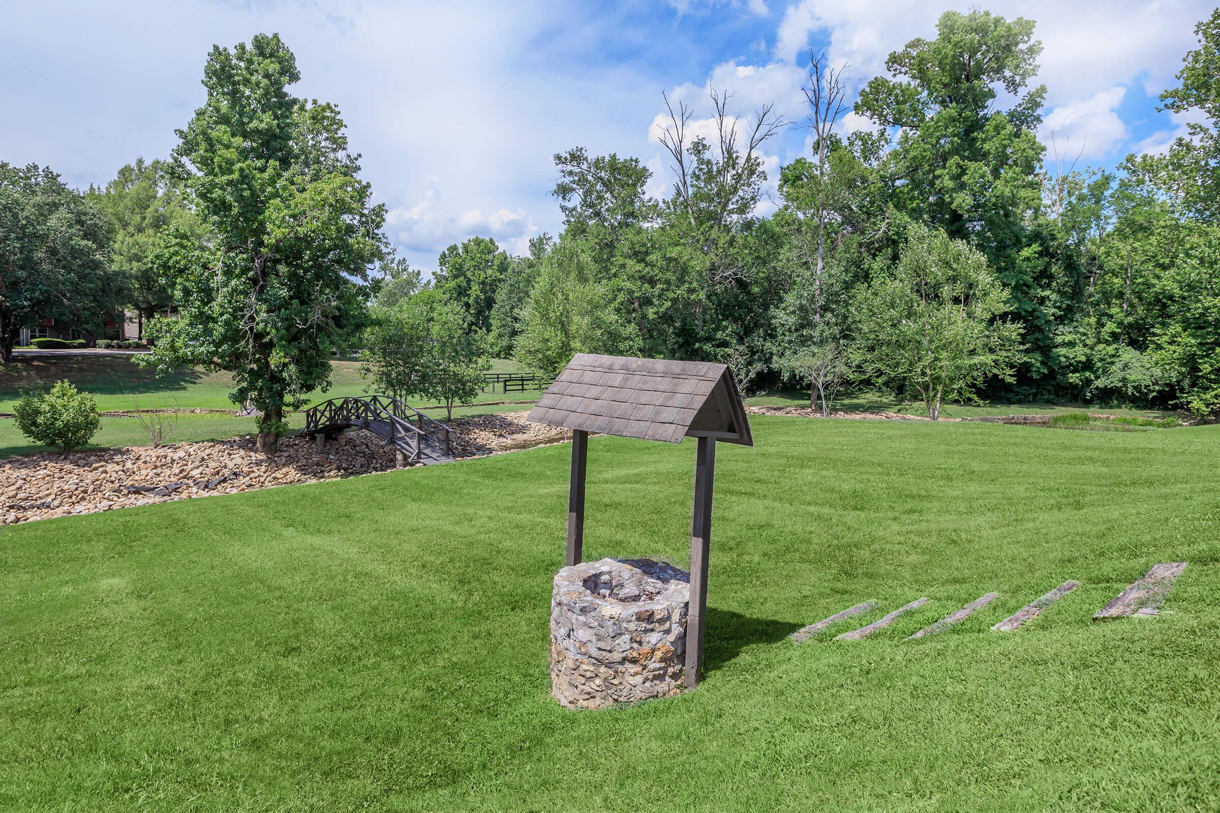 A stone well with a wooden roof situated in a lush, green yard surrounded by trees. The scene includes a wooden bridge in the background and a grassy area with a few stone steps leading away from the well, under a partly cloudy sky.