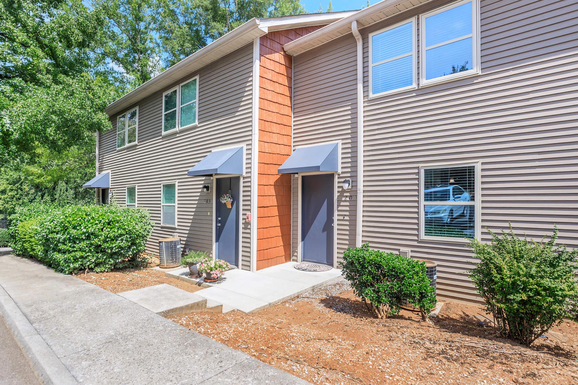 Two side-by-side townhome entrances with gray doors and awnings, surrounded by small shrubs and mulch. The building has beige siding with a reddish-orange accent on one side. The area is well-lit by sunlight and there are trees in the background, providing a vibrant, welcoming atmosphere.