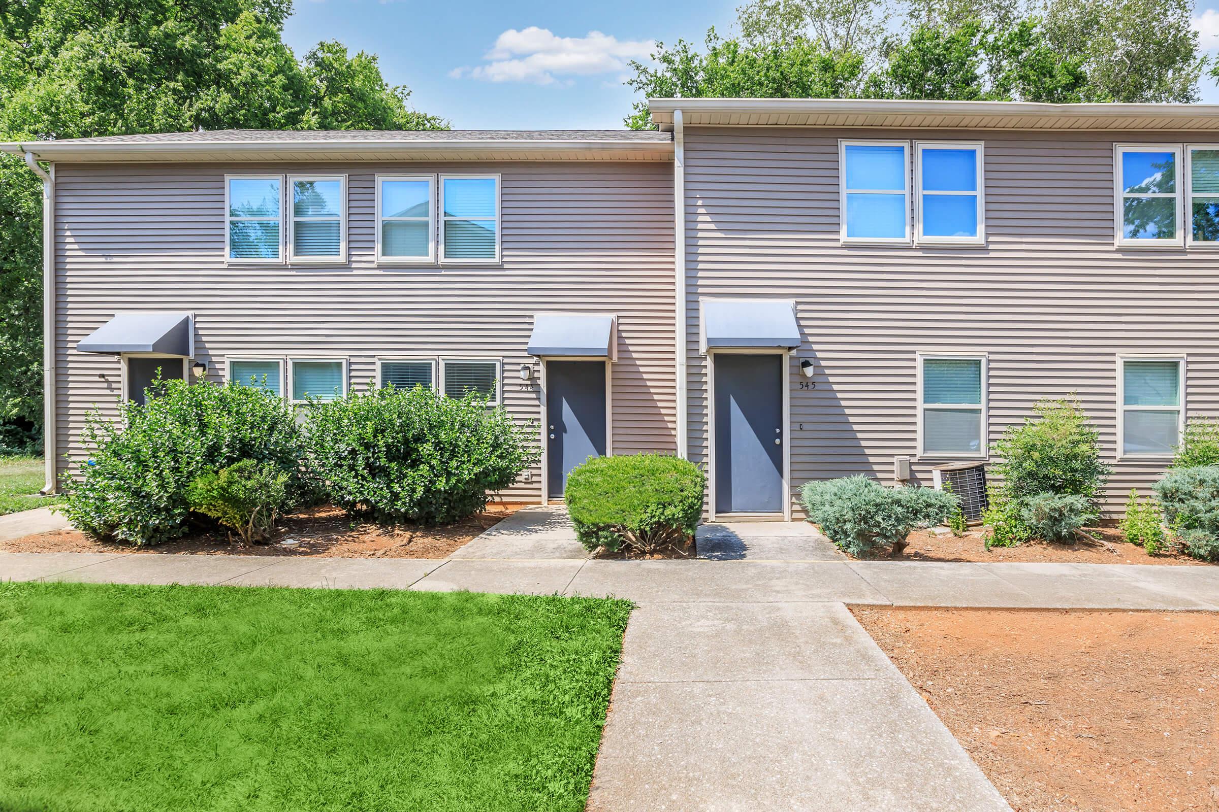 A view of a pair of residential buildings, showcasing two entryways with dark doors and blue awnings. The surrounding area features green shrubs and a well-maintained lawn. A clear blue sky is visible above, contributing to a bright and welcoming atmosphere.