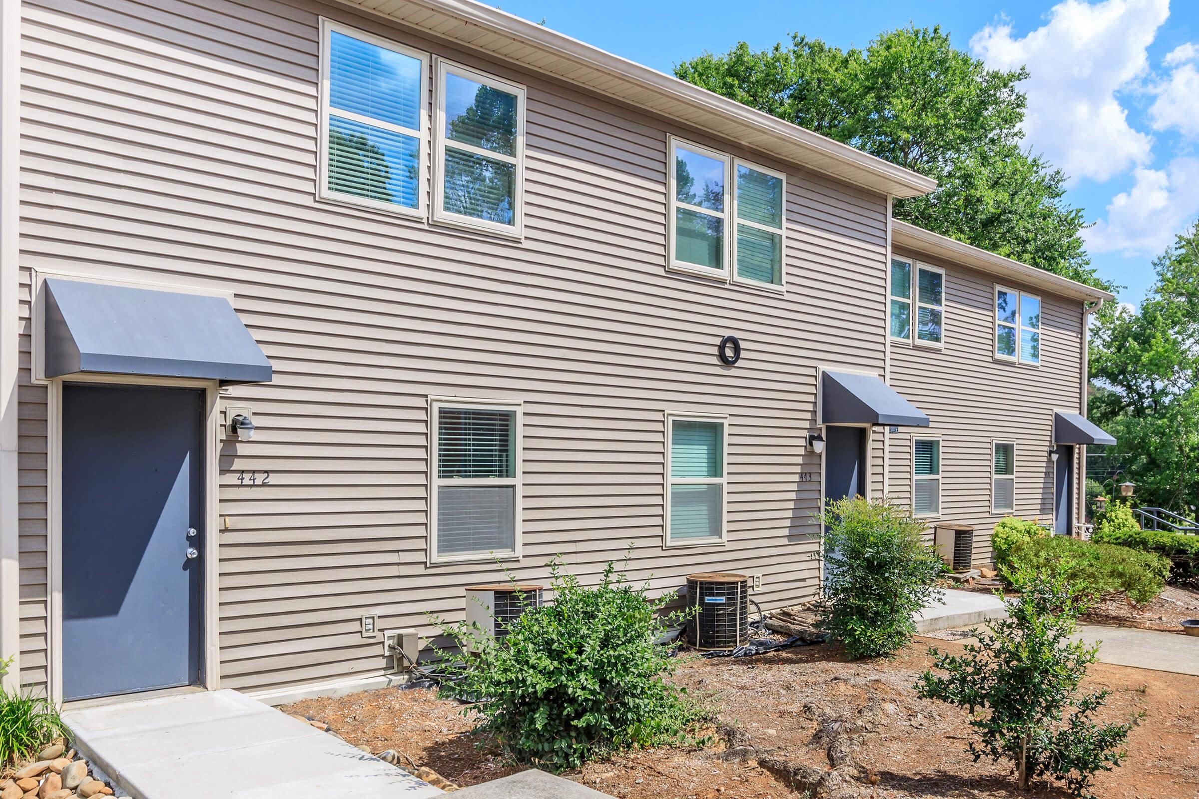A row of beige-colored townhouses with gray doors and awnings. Each unit has windows with white frames and is surrounded by small shrubs and landscaping. The pathway in front is made of concrete, and there are patches of mulch and grass in the yard, with a clear blue sky above.