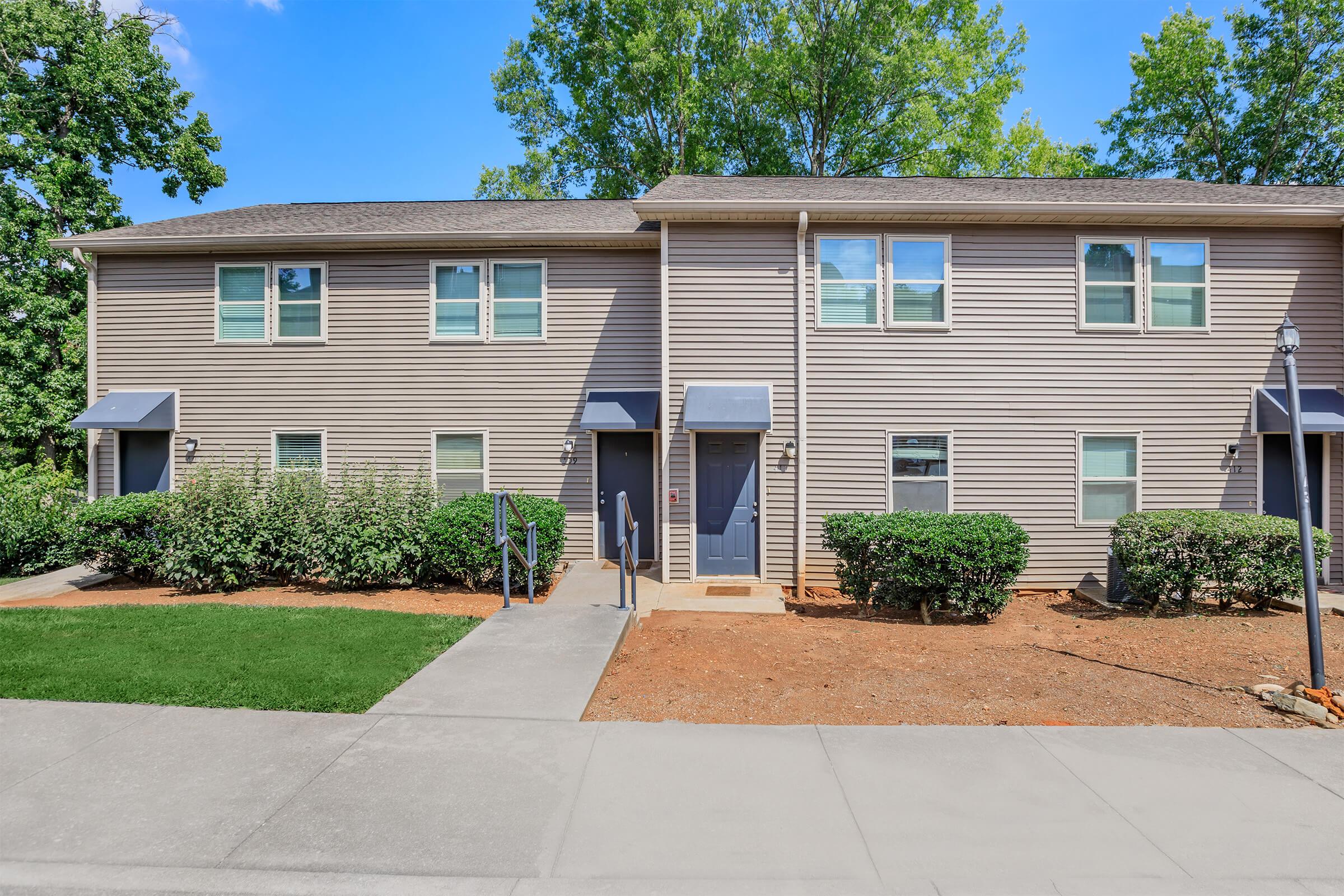 A view of a two-story apartment building with light brown siding, featuring multiple windows and blue front doors. The pathway leads to the entrance, bordered by manicured bushes and grass. Trees are visible in the background under a clear blue sky.