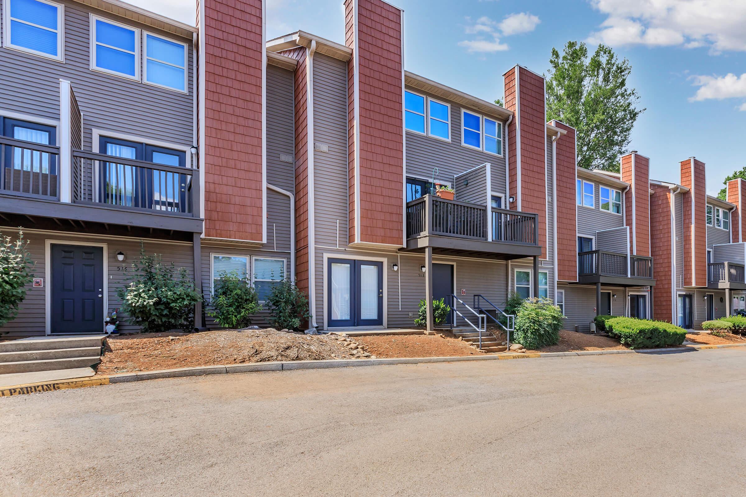 Row of modern townhouses with red and gray siding, balconies on the upper levels, and greenery in front. The street is paved with a clear blue sky above. Steps lead up to the door of one unit, and parking is visible along the side.