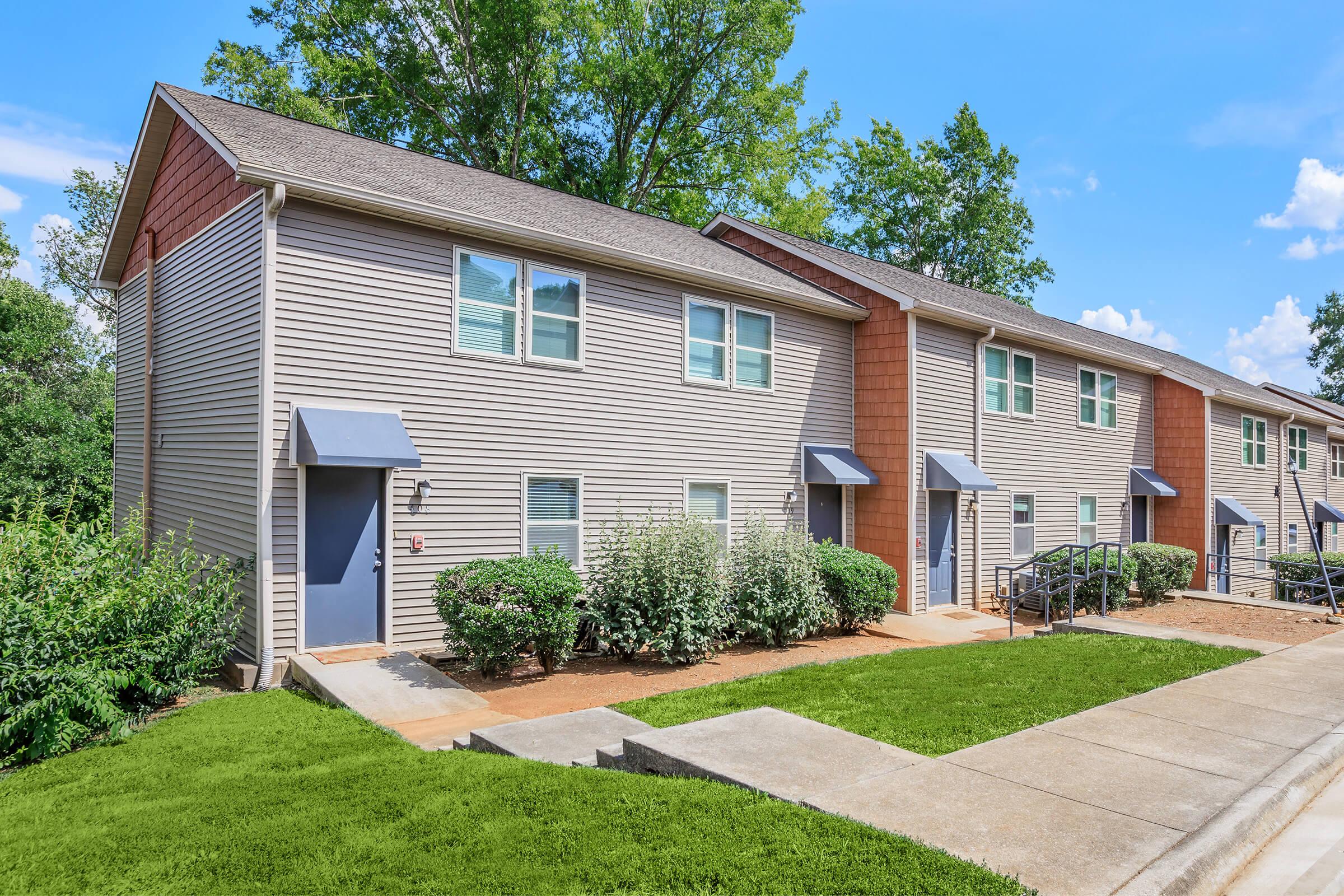 A row of townhouse-style apartments with gray siding and brown accents. Each unit features a door with a small porch and window. Surrounding greenery includes bushes and trees, set against a clear blue sky with scattered clouds. A concrete path leads to the entrance of each home.