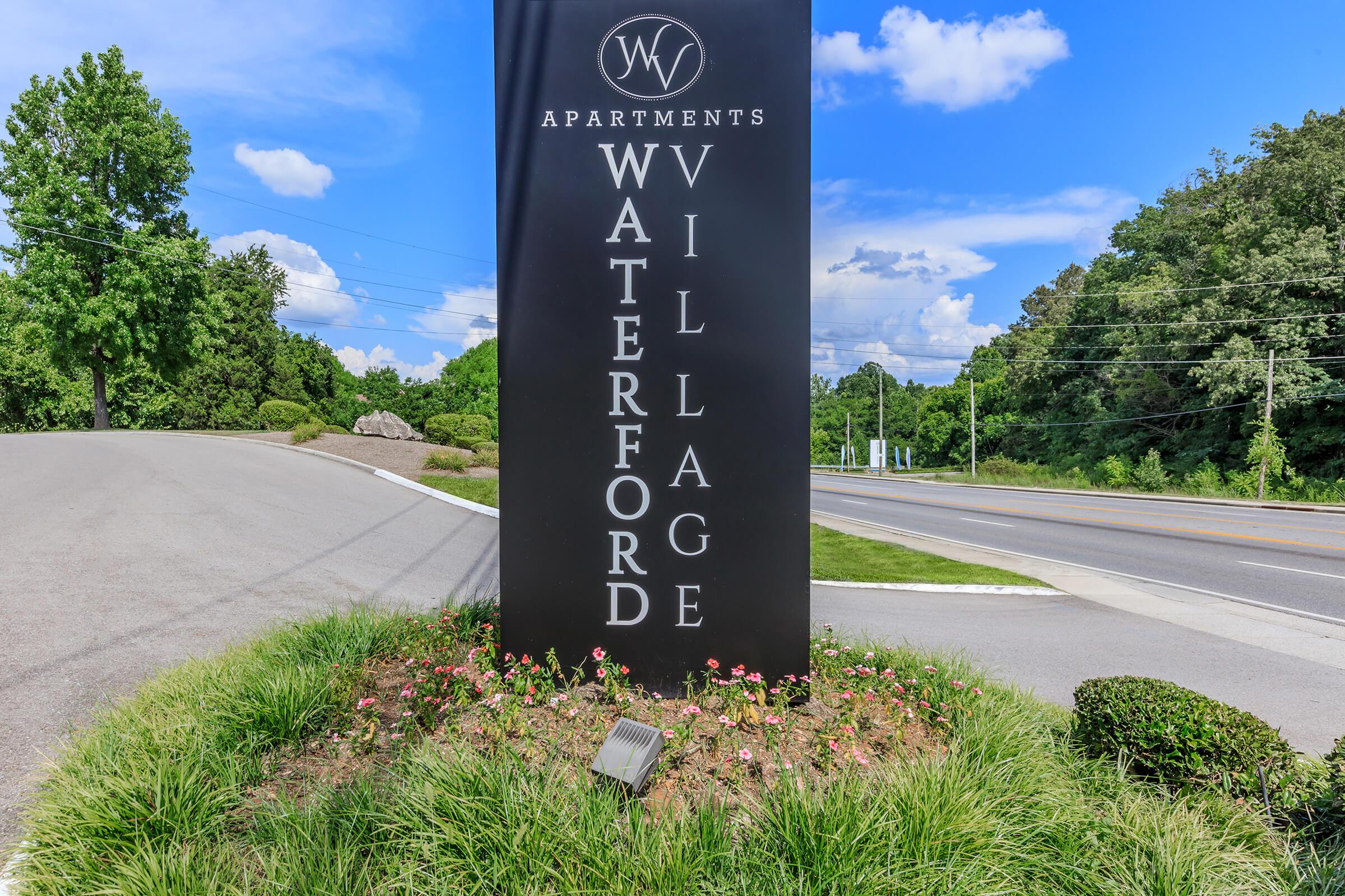Sign for Waterford Village Apartments, featuring a sleek black design with the text "Waterford Village" prominently displayed. Surrounding the sign are landscaped areas with green grass and small flowers, set against a backdrop of blue sky and trees. The sign is positioned near a road.