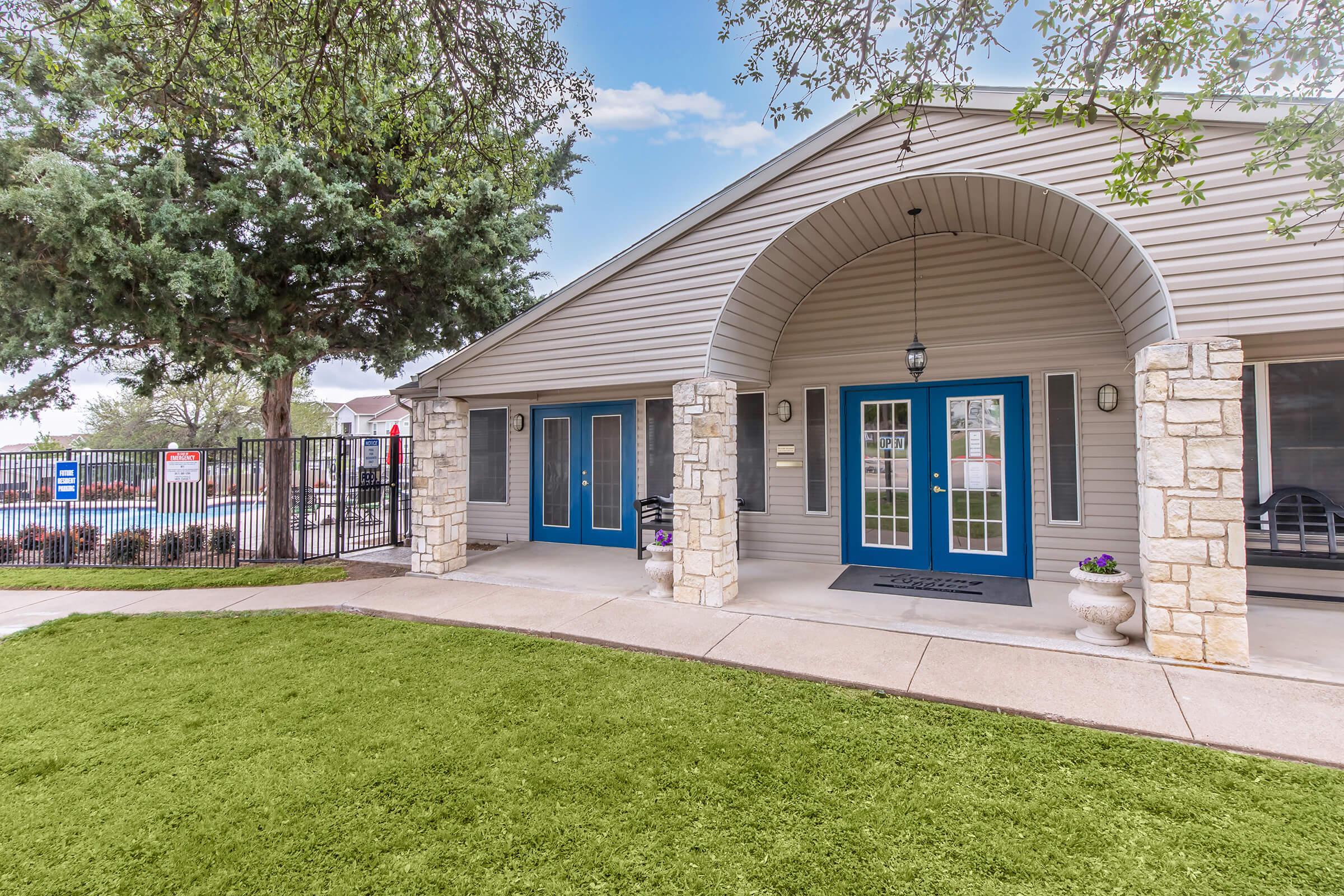 Entrance to a modern building featuring a curved archway, blue double doors, and stone accents. The pathway is bordered by green lawn, with potted plants near the entrance. In the background, a fenced area and trees are visible under a clear blue sky.