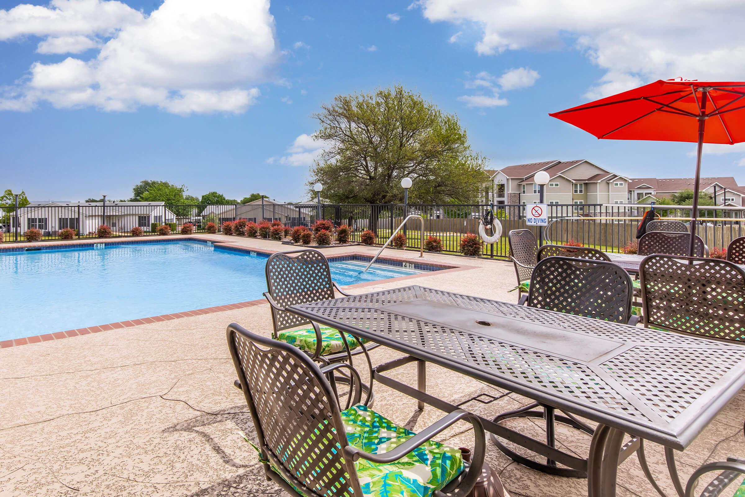 A clear blue swimming pool surrounded by a patio area with metal tables and chairs. An orange umbrella provides shade by the poolside. Lush greenery and flowering plants are visible along the fence. Soft clouds float in a bright blue sky, creating a pleasant outdoor atmosphere.