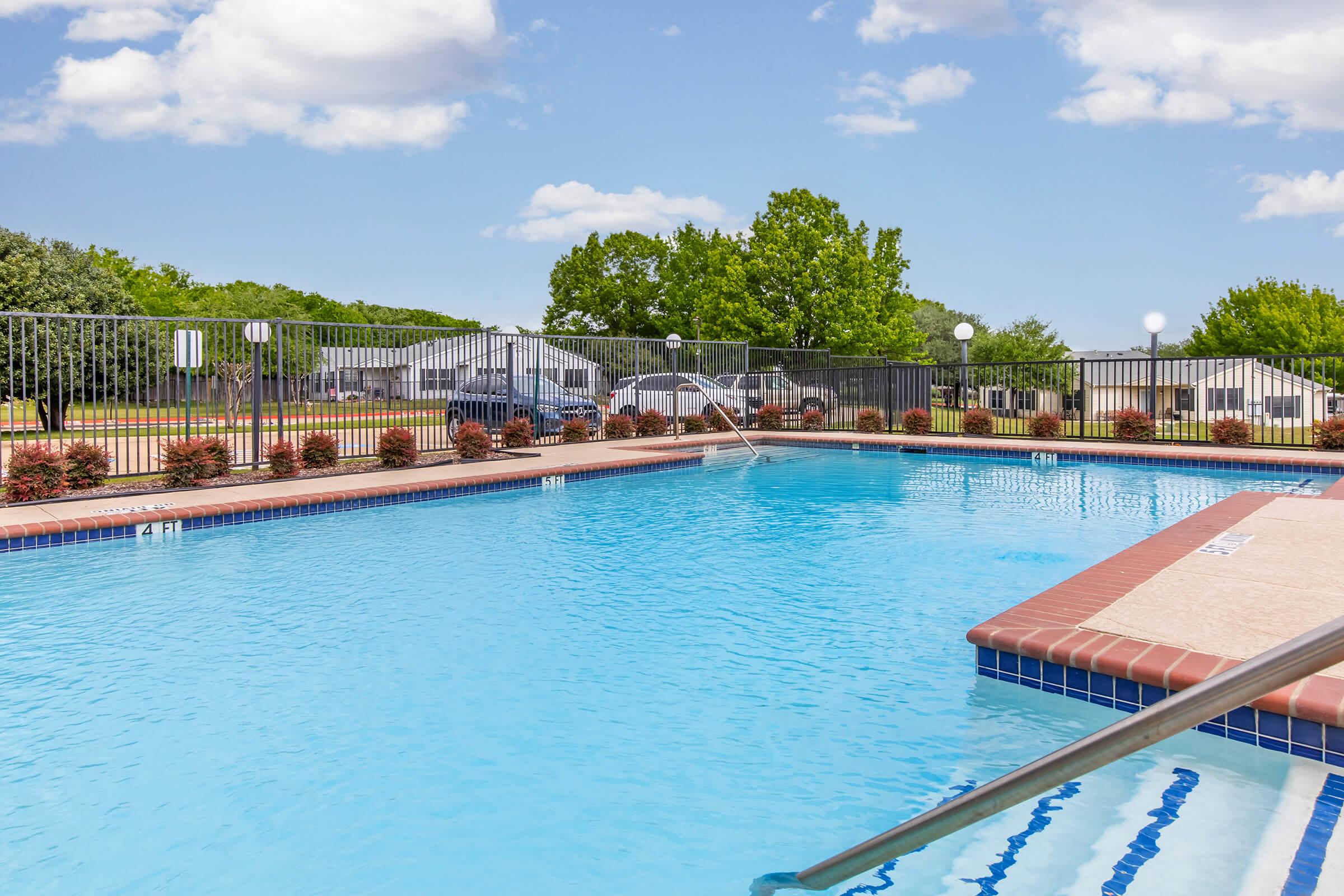 A clear blue swimming pool surrounded by a wrought-iron fence, featuring steps leading into the water. Lush green trees and a bright sky with fluffy clouds are visible in the background, along with a few parked vehicles and landscaped shrubs along the pool's edge.