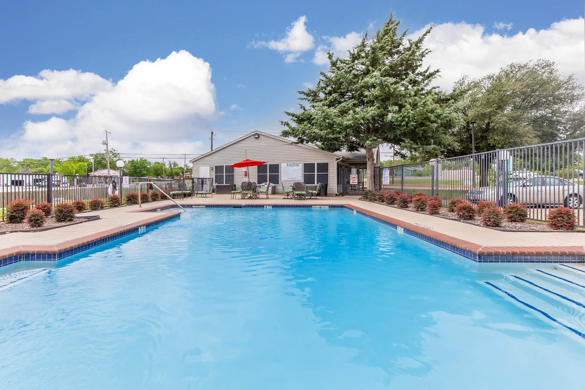 Swimming pool surrounded by landscaped areas, featuring lounge chairs and a red umbrella. A building is visible in the background, with a fenced area and clear blue sky. The scene exudes a calm and inviting atmosphere, perfect for relaxation.