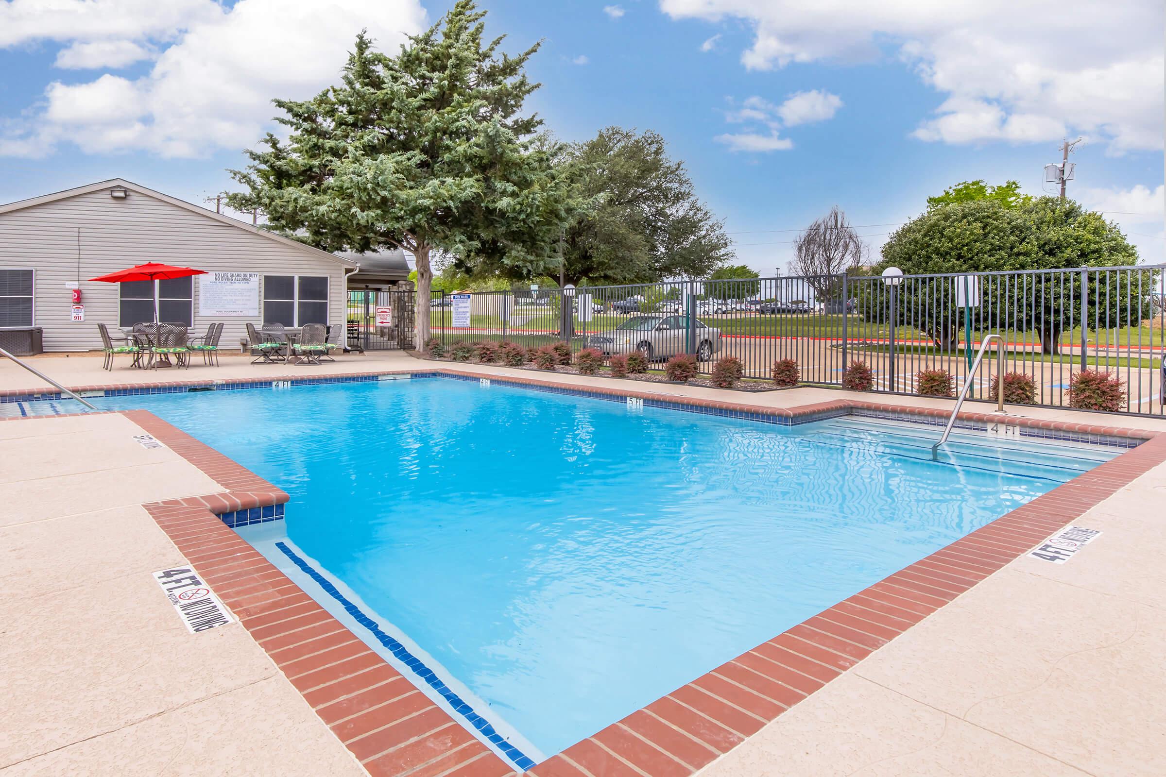 A well-maintained outdoor swimming pool surrounded by a landscaped area. The pool features clear blue water, a ladder for entry, and a patio with tables and chairs, as well as a shade umbrella. In the background, there is a building and various trees, with a blue sky and fluffy clouds above.