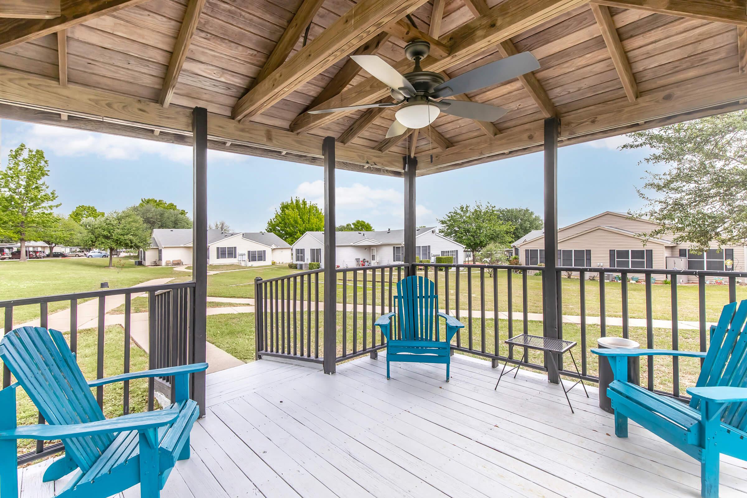 A spacious outdoor gazebo featuring wooden beams and a ceiling fan. Inside, there are two turquoise Adirondack chairs and a small table, with green grass and houses visible in the background under a clear blue sky.