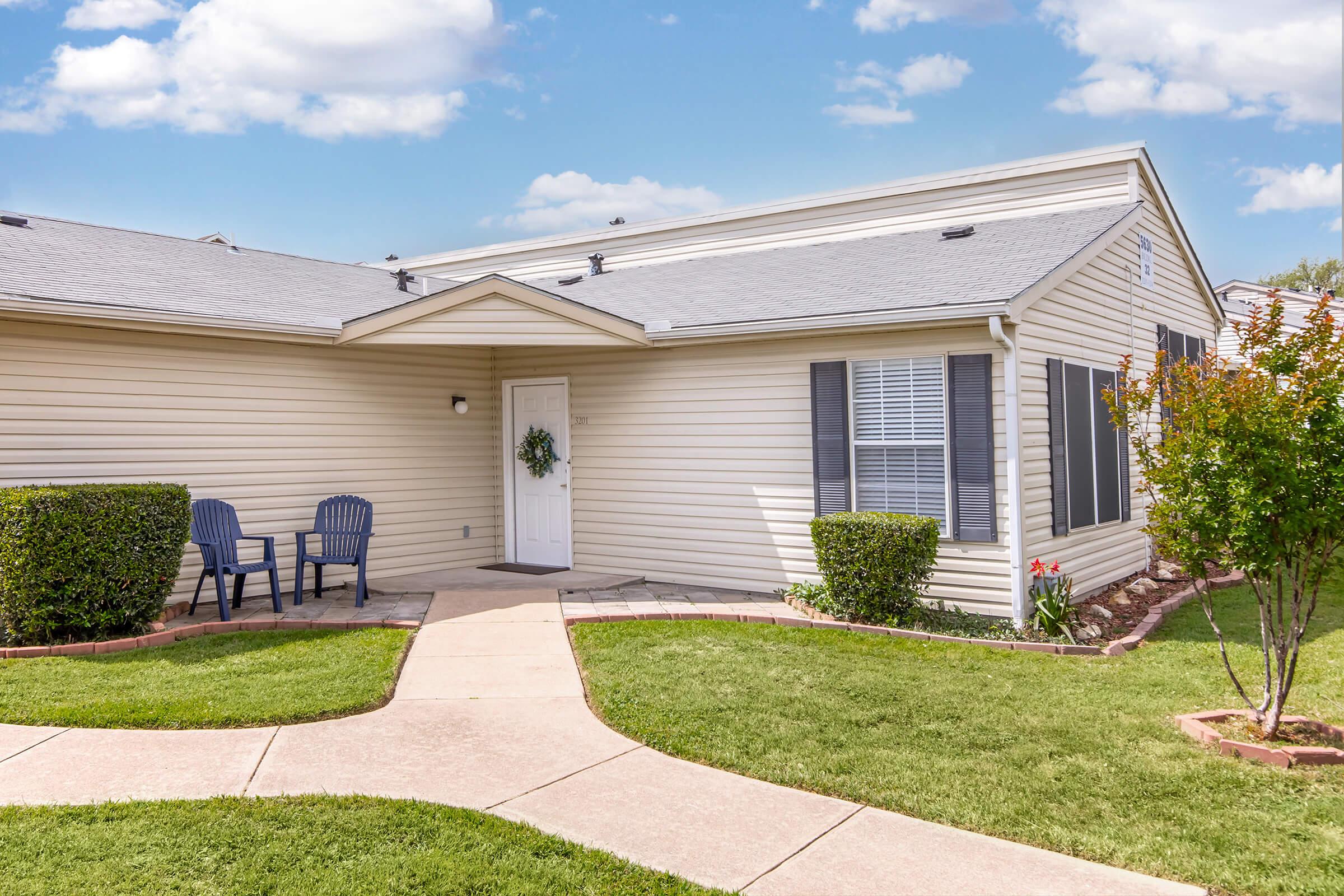 A neatly landscaped exterior of a single-story home with light-colored siding. The entrance features a white door adorned with a wreath. There are two dark chairs on the porch, and a well-maintained pathway leads to the door, bordered by green grass and decorative shrubs. A clear blue sky is visible in the background.