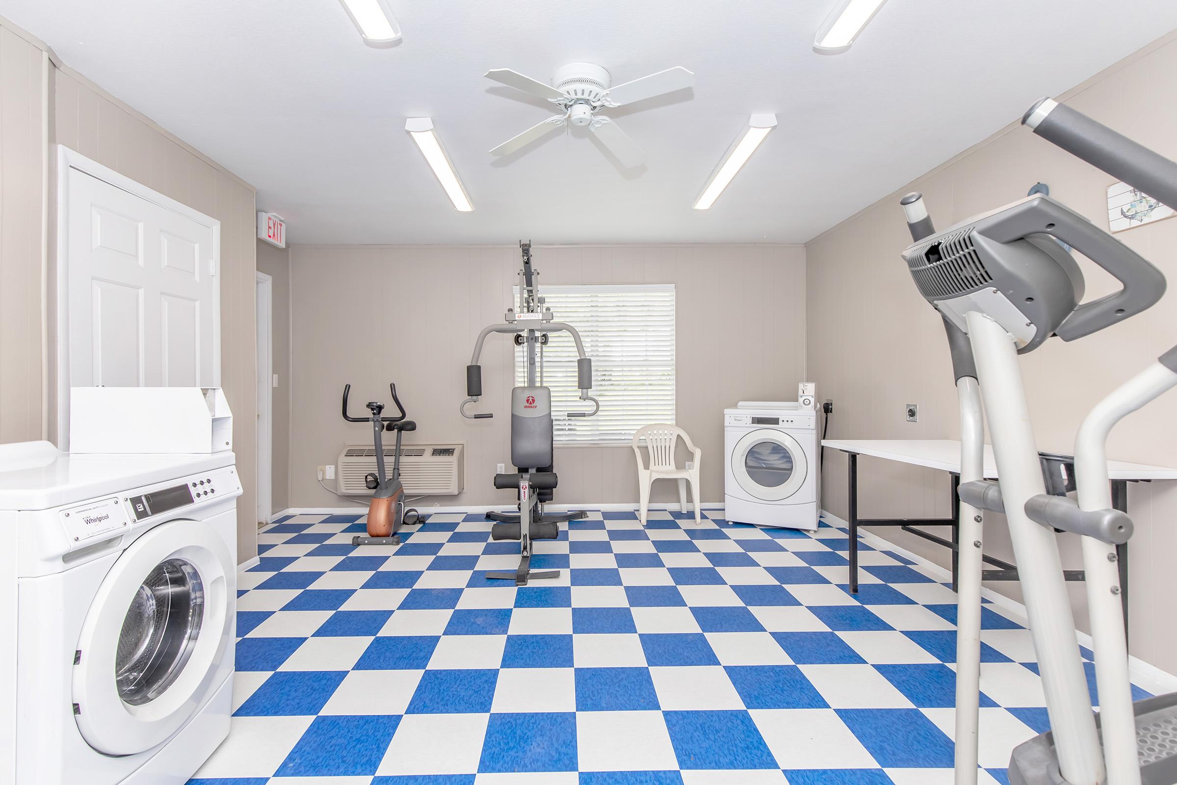 A bright gym or laundry room featuring workout equipment like a multi-gym machine and an exercise bike, along with a washing machine and dryer. The room has a blue and white checkered floor, a ceiling fan, and large windows providing natural light.