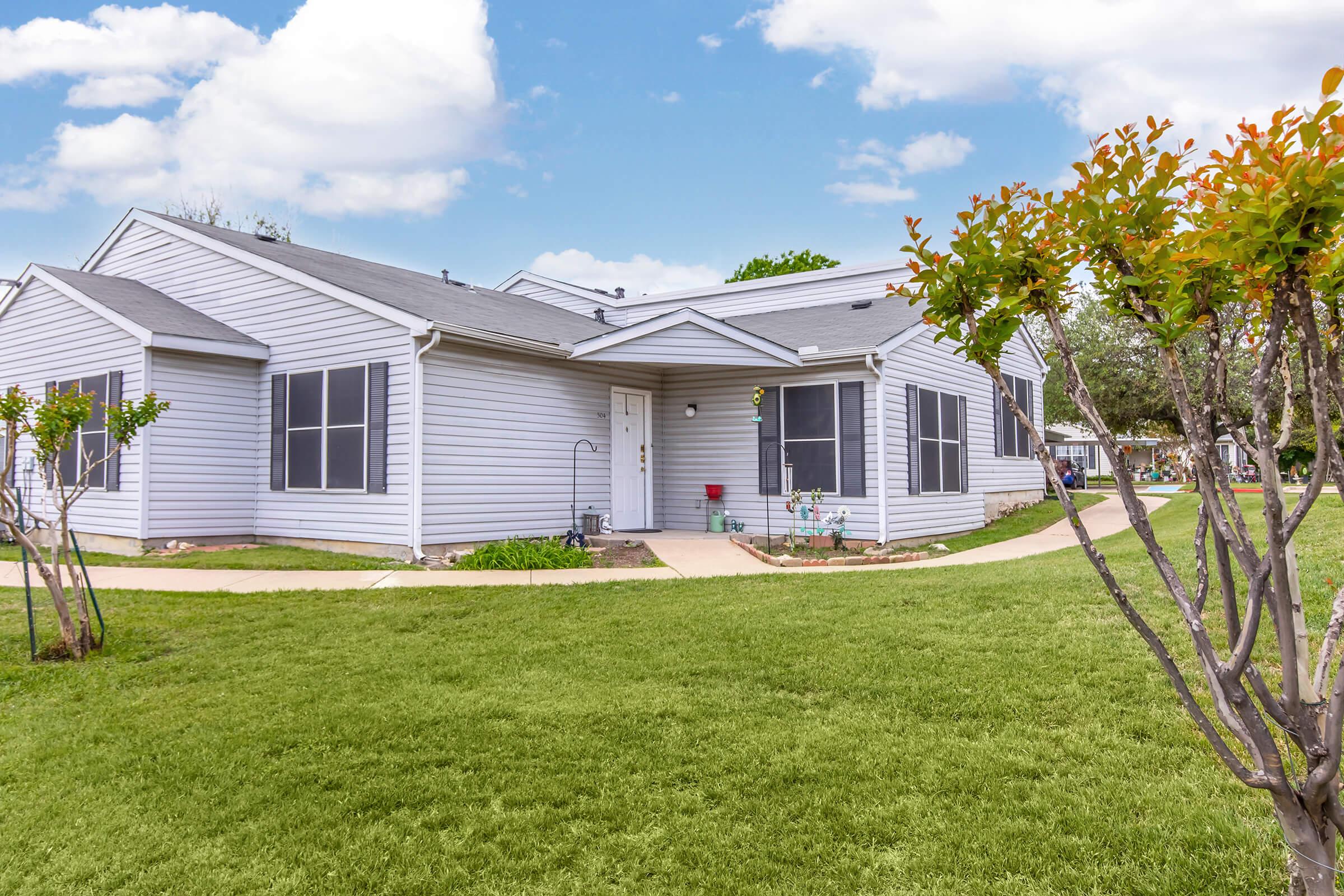 A modern single-story home with a light gray exterior and multiple windows, surrounded by well-maintained grass and a pathway. In the foreground, there are small shrubs, and the sky above features fluffy white clouds. The overall setting appears bright and inviting.