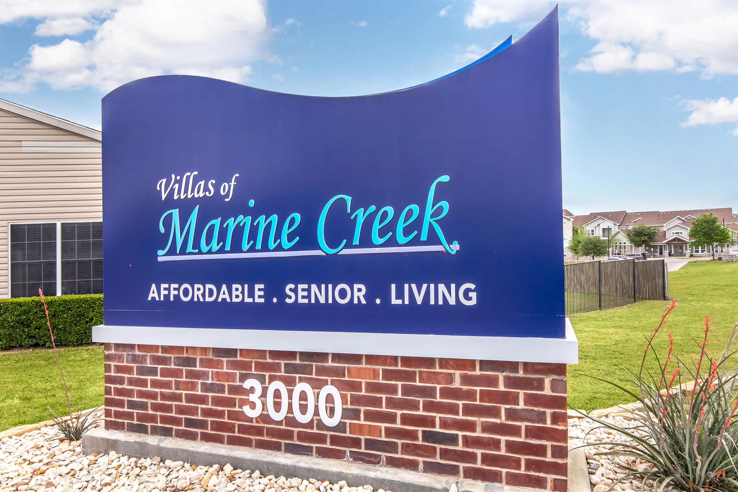 Sign for "Villas of Marine Creek" indicating "Affordable Senior Living," situated on a brick base with a decorative landscape. The sign features a blue background with white and teal lettering, set against a clear sky and a green lawn. The location number "3000" is displayed prominently at the bottom.