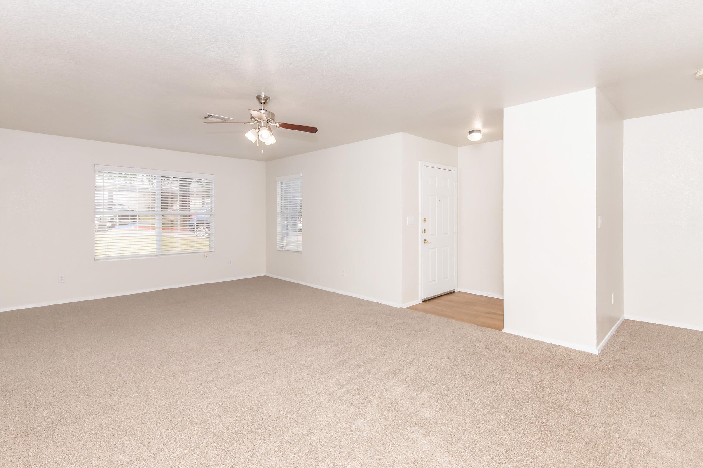 A spacious, empty living room with light beige carpet and white walls. A ceiling fan is installed, and there are two windows letting in natural light. To the right, a white door leads outside. A dividing wall creates a separate entry space. The overall atmosphere is bright and inviting.