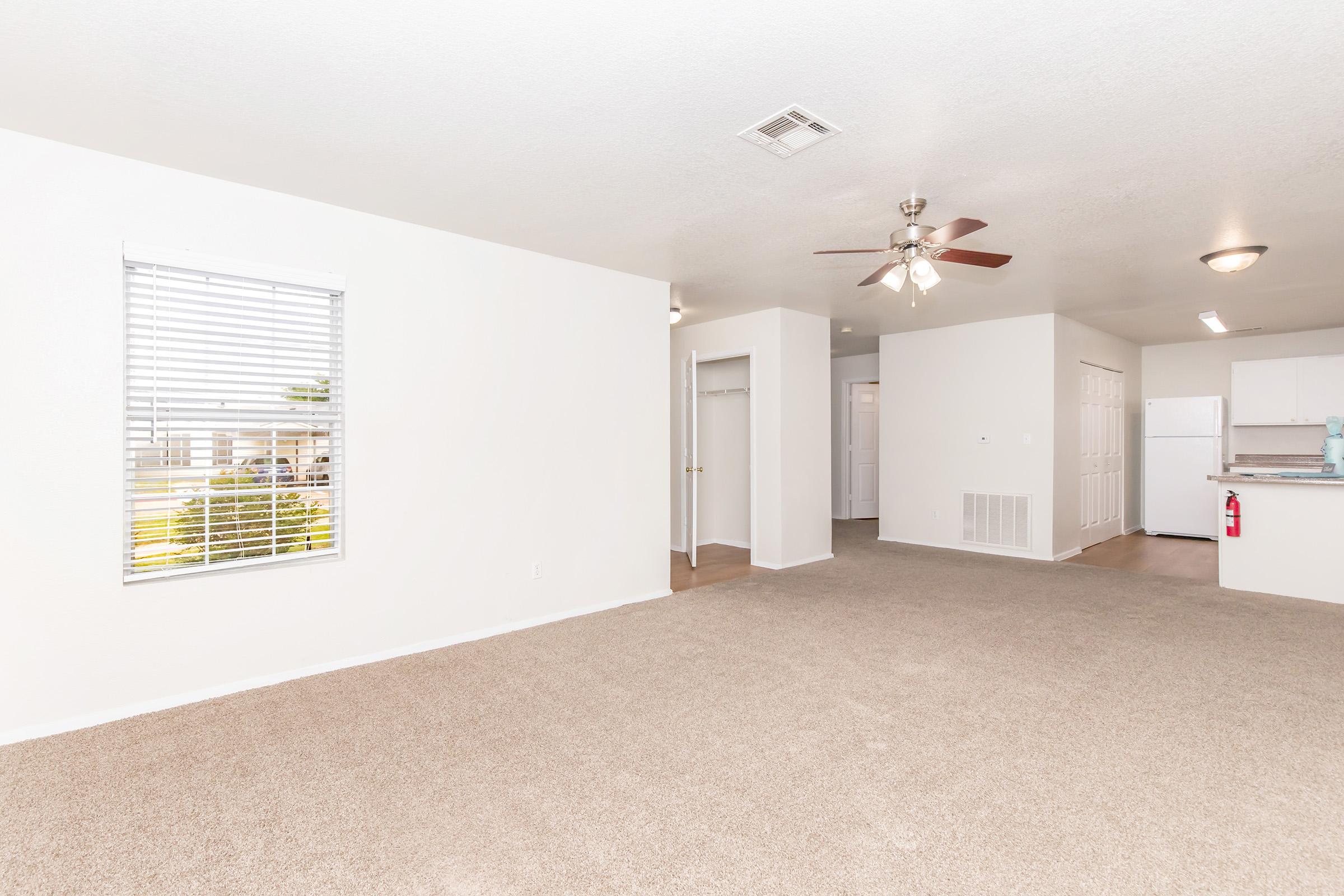 A spacious, well-lit interior of an empty apartment featuring beige carpet, white walls, and a ceiling fan. A window with blinds allows natural light, and there's an open layout connecting to a kitchen area with white appliances. The overall atmosphere is fresh and welcoming.