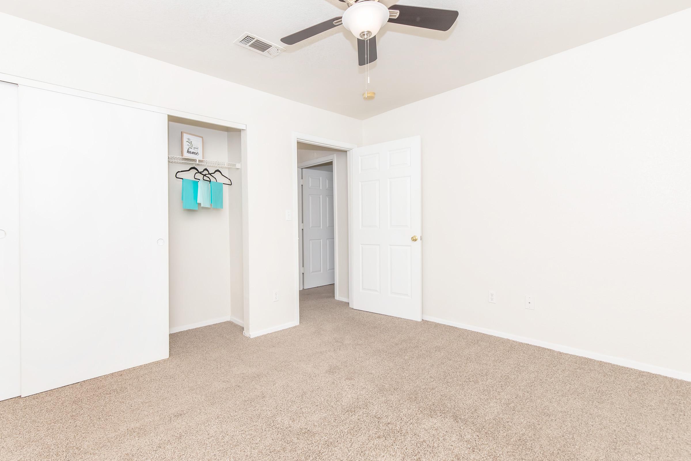 A view of a well-lit room with beige carpet, featuring a ceiling fan. To the left, there is a closet with white sliding doors and two hanging items. An open door leads to another room, and the walls are painted a light color, creating a bright and spacious atmosphere.