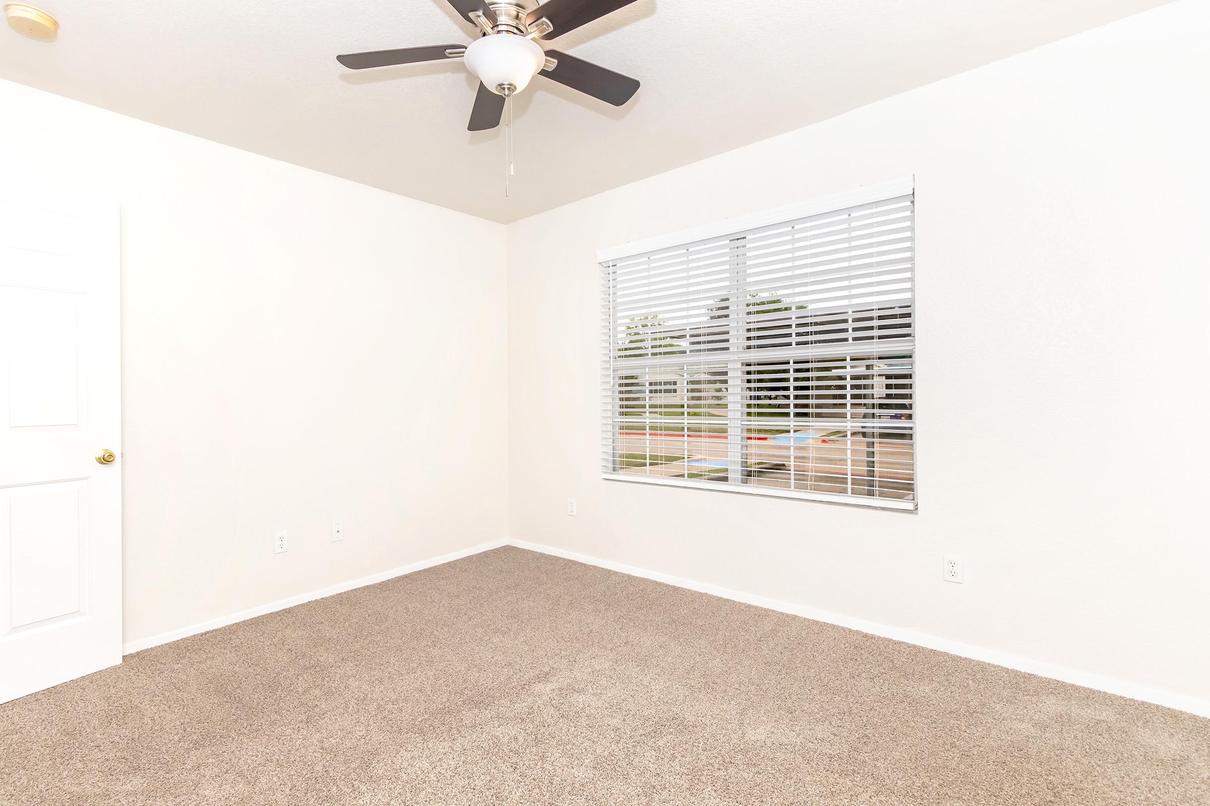 A clean, empty room featuring a light-colored wall and carpet. A ceiling fan hangs from the ceiling, and there’s a window with white blinds allowing natural light to enter. The room appears bright and spacious, ready for furnishing.