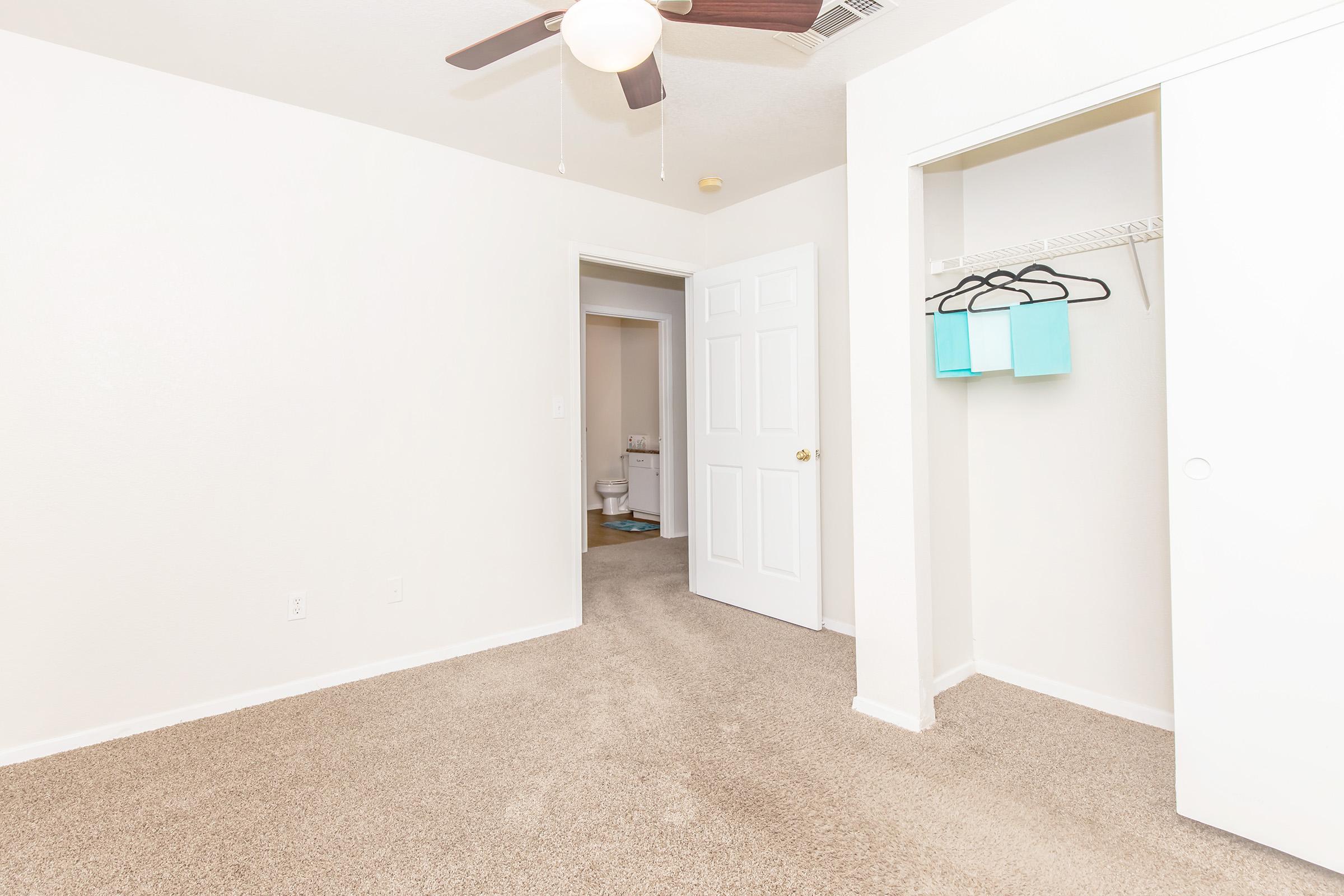 A bright, empty bedroom featuring light-colored walls and carpet. There is a ceiling fan and an open closet with blue hangers visible. A door leads to a bathroom in the background, which is partially visible. The room feels spacious and well-lit.