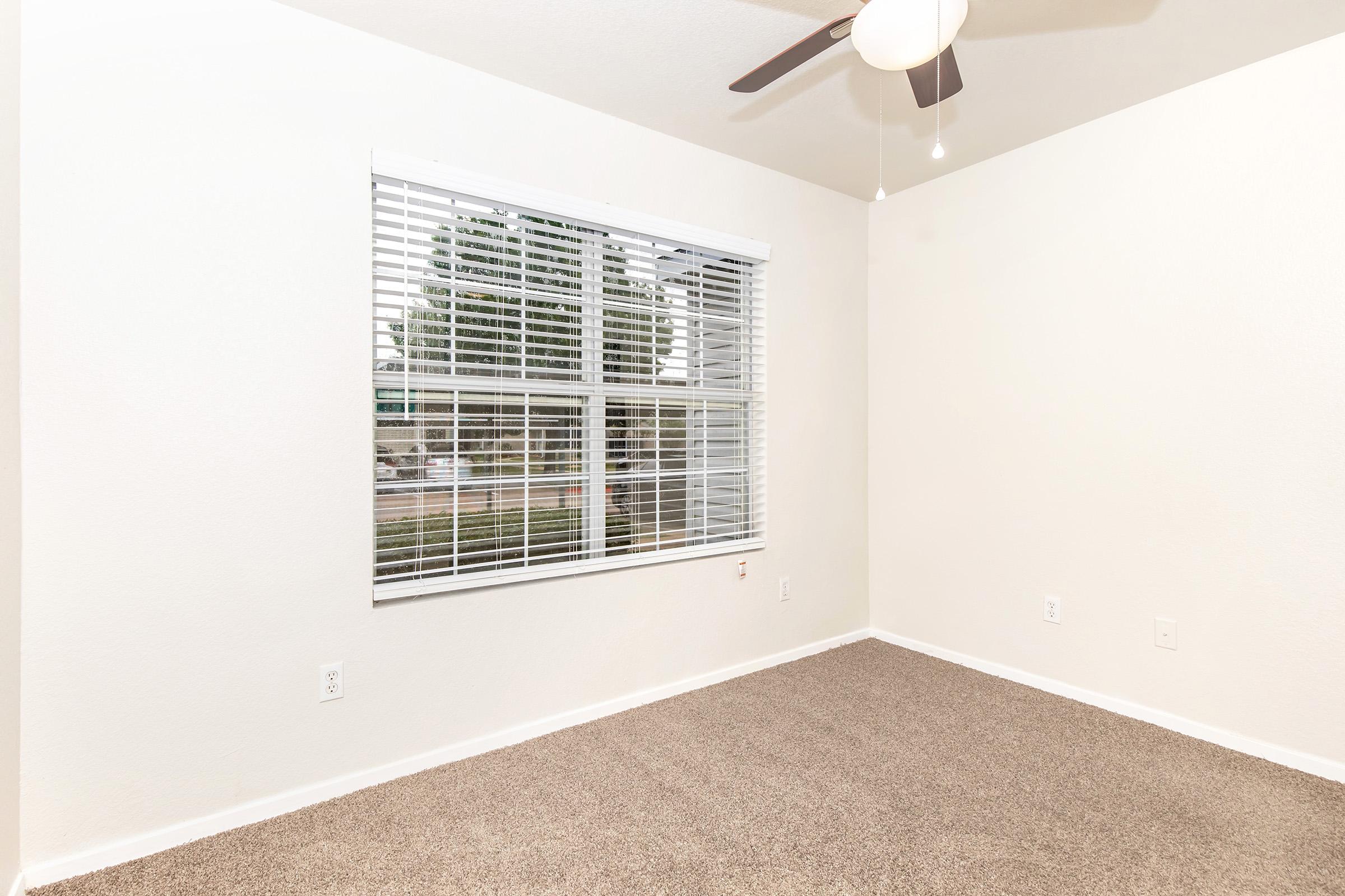 An empty room with light beige walls and soft carpet flooring. A large window with white blinds lets in natural light, illuminating the space. A ceiling fan is visible, contributing to the modern aesthetic of the room. The overall atmosphere is clean and minimalistic.