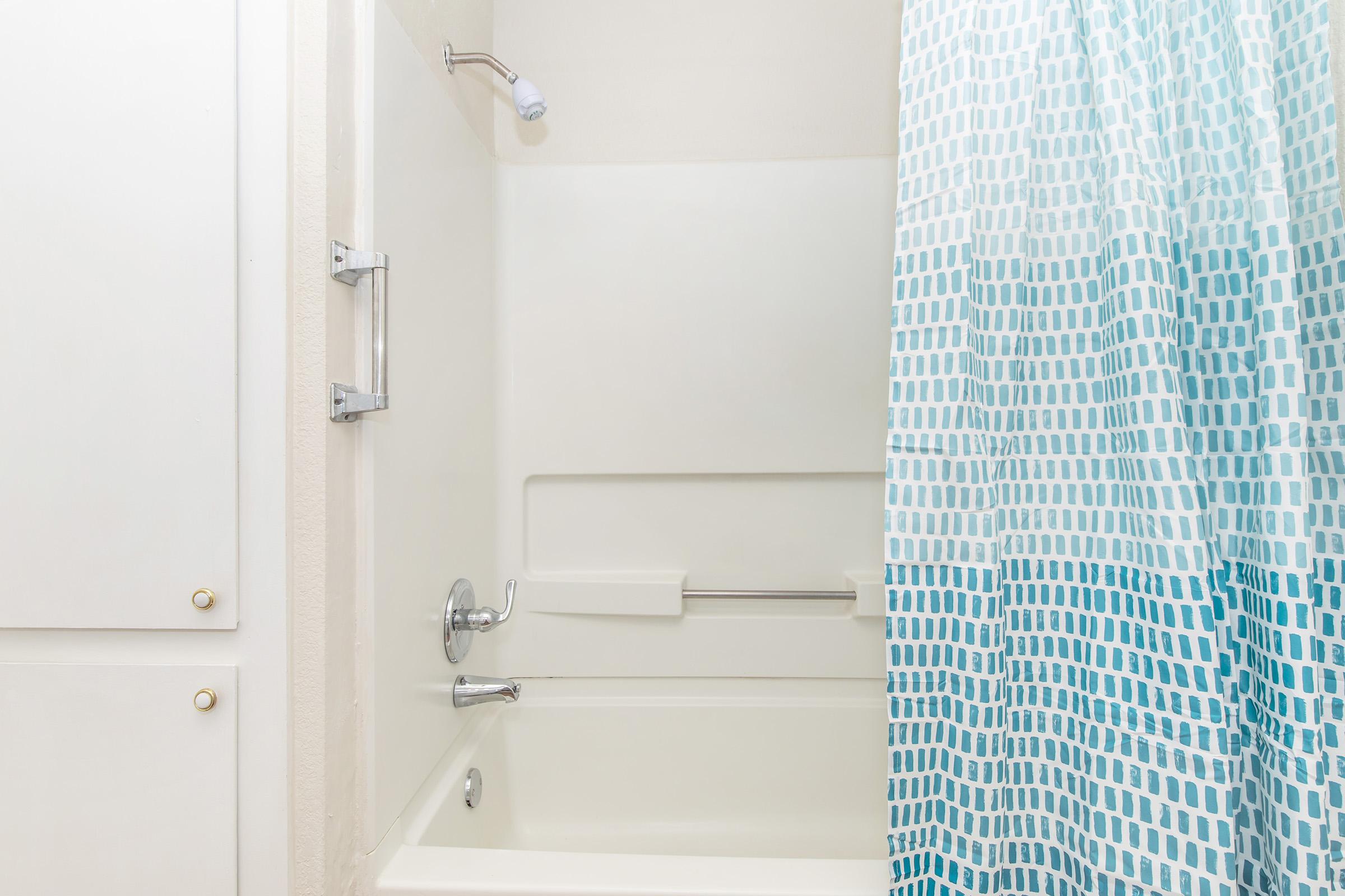A clean bathroom with a white bathtub and a light blue patterned shower curtain. The shower area features a showerhead and a grab bar. There is a white cabinet or closet beside the bathtub, providing storage space. The overall ambiance is bright and fresh.