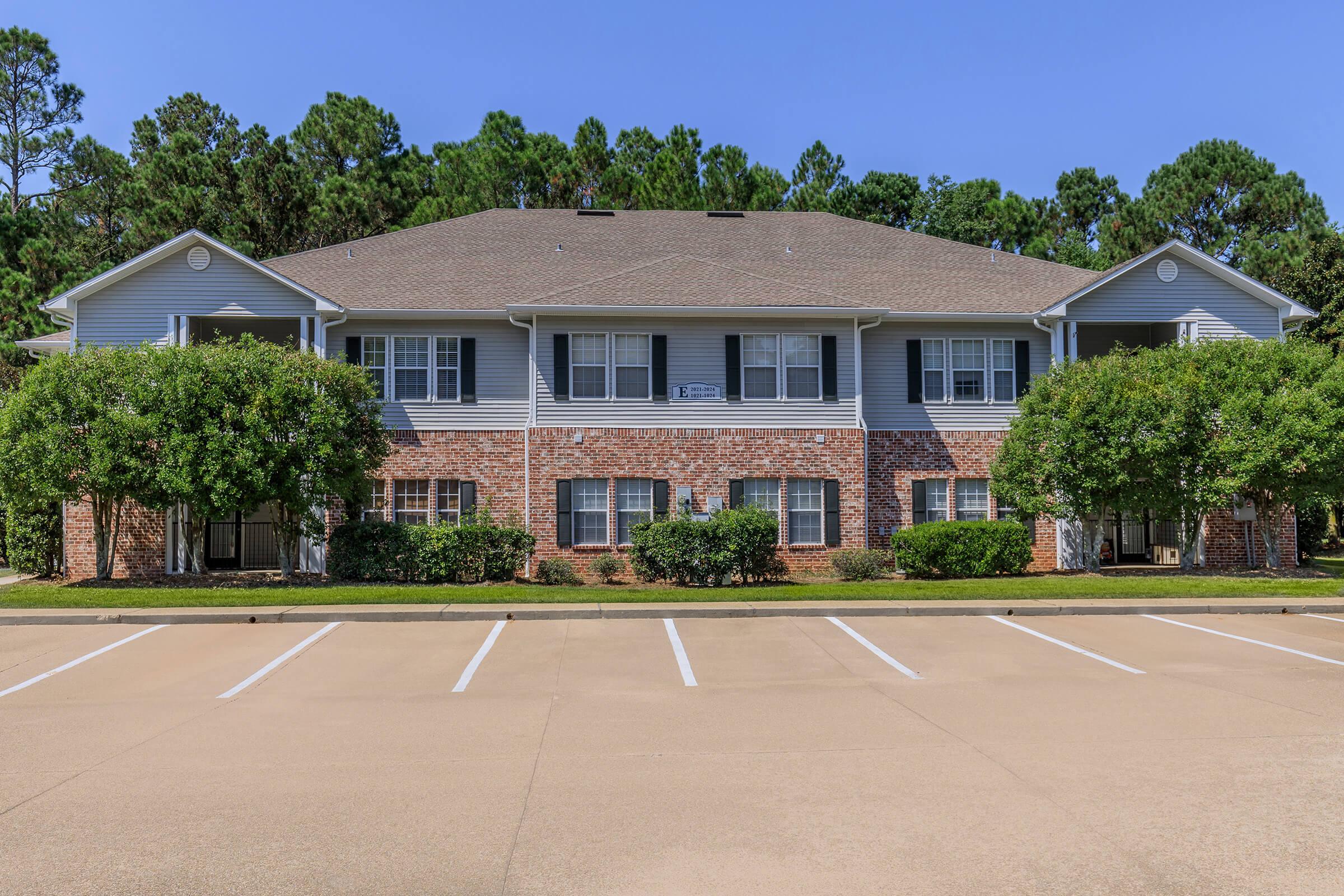 A multi-unit residential building with a brick and siding exterior, featuring multiple balconies and large windows. The landscaping includes green shrubs and trees, with a paved parking lot in front containing empty parking spaces under a clear blue sky.