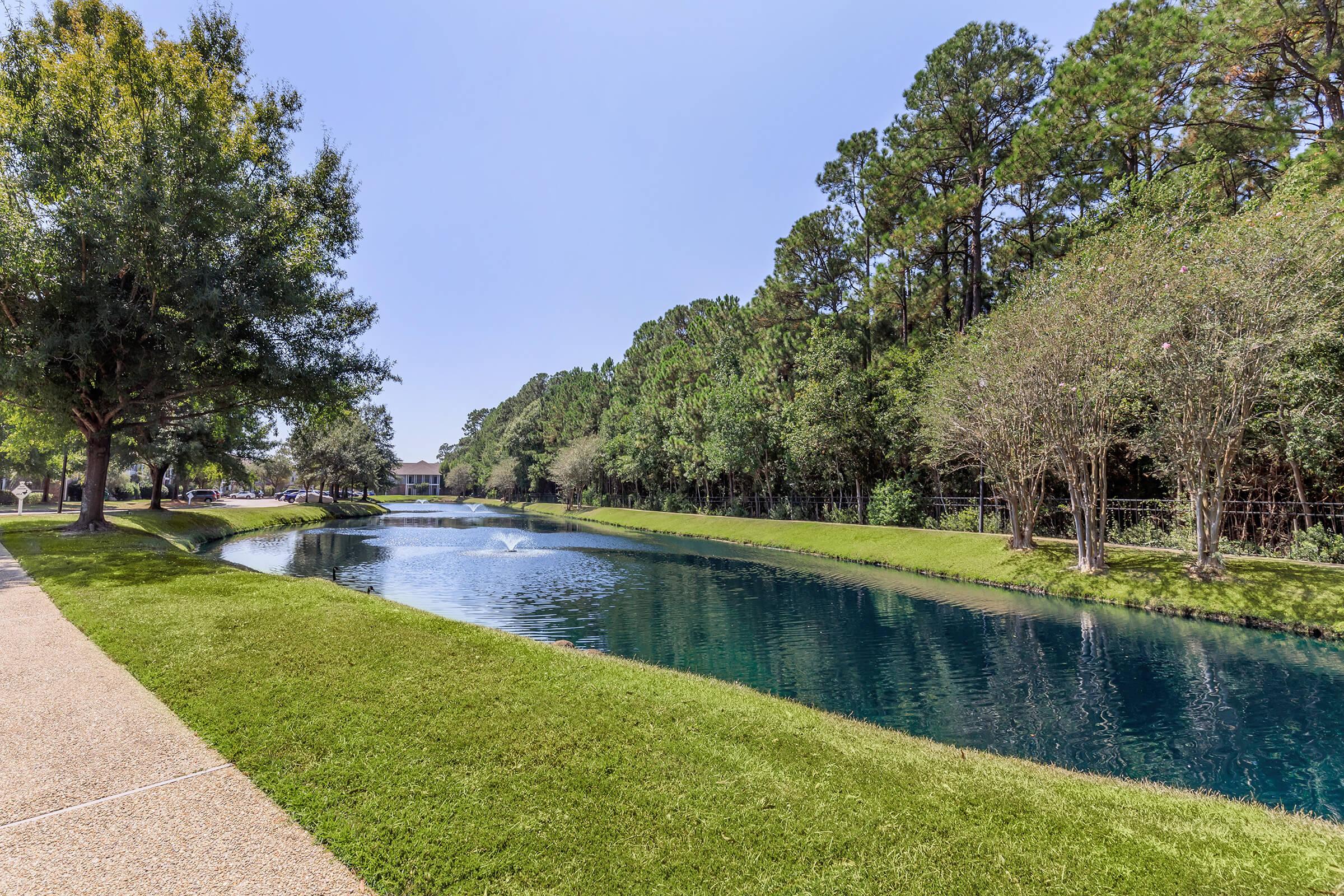 A serene landscape featuring a calm pond surrounded by lush green grass and trees. A gentle fountain can be seen in the water, reflecting the clear blue sky and creating a tranquil atmosphere, perfect for a peaceful stroll along the paved walkway.