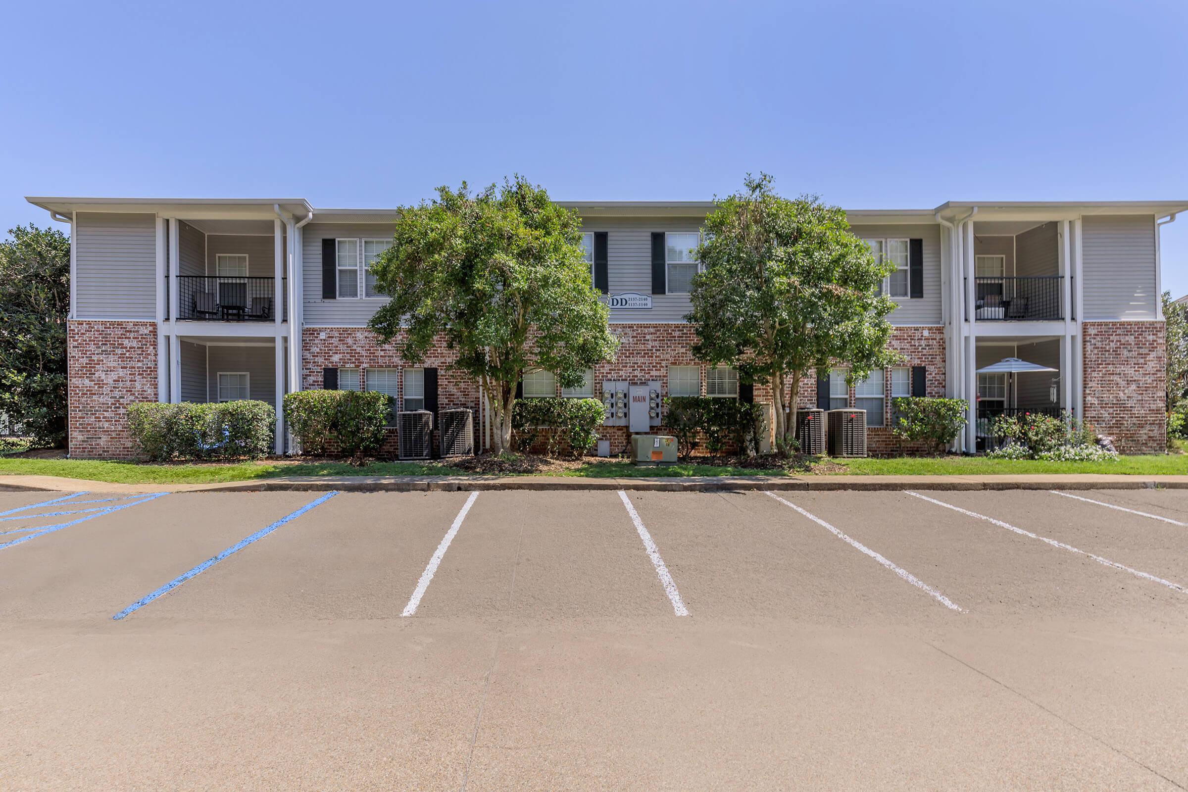 A two-story apartment building with brick and siding exterior. The building features balconies with green shrubs and trees in front. A parking lot is visible with empty parking spaces marked in blue. The sky is clear and sunny, providing bright lighting to the scene.