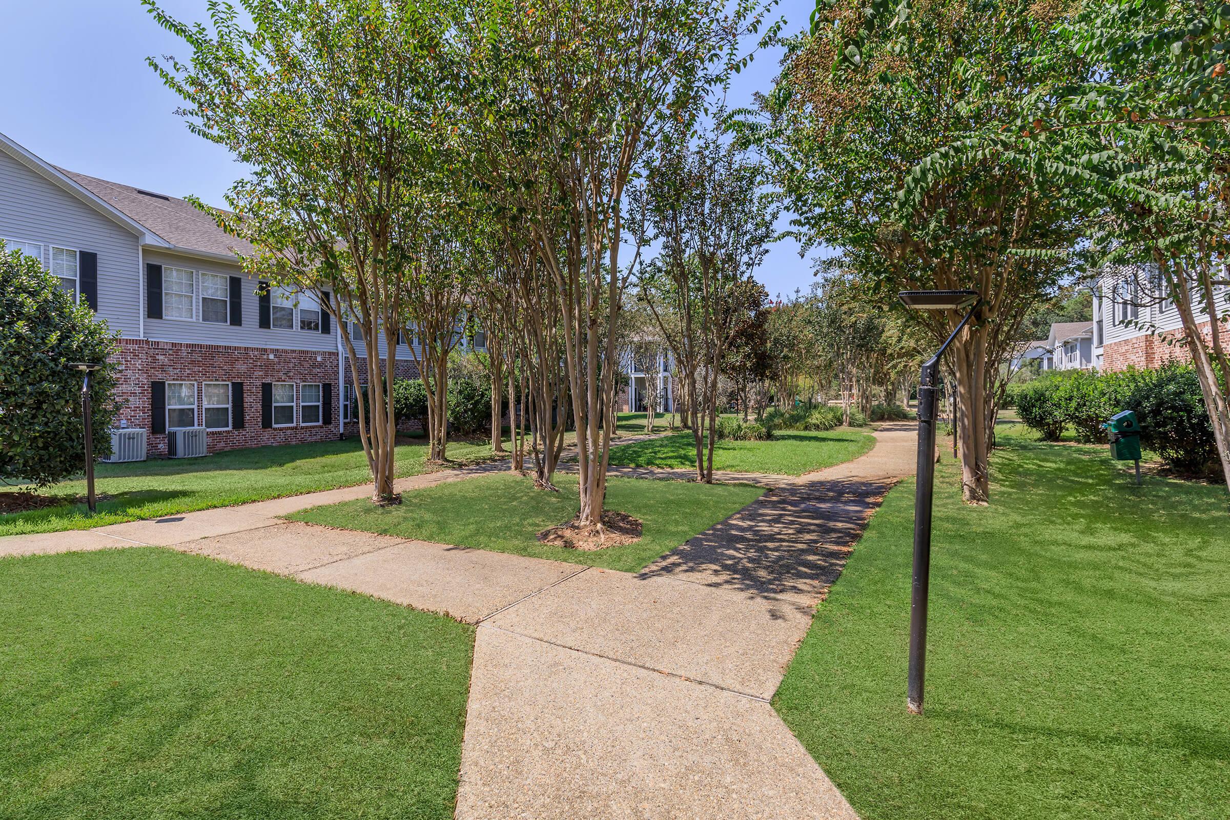 A landscaped outdoor area featuring walking paths winding through well-maintained green grass, bordered by small trees and shrubs. The scene includes a multi-story building in the background, with several windows visible, and decorative lighting fixtures along the paths. Clear blue sky overhead.