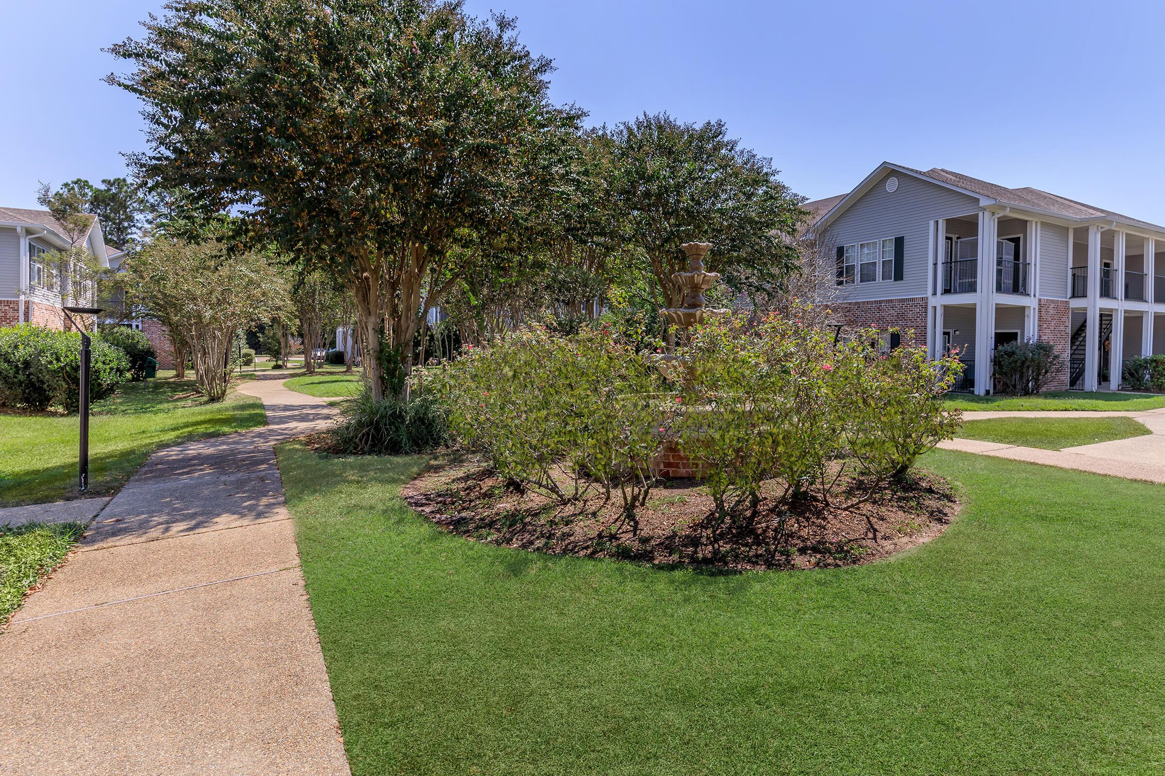 A well-maintained landscape featuring a circular garden bed with a small fountain in the center, surrounded by shrubs. Sidewalks lead through the area, with buildings visible in the background under a clear blue sky. The scene exudes a peaceful and inviting atmosphere.