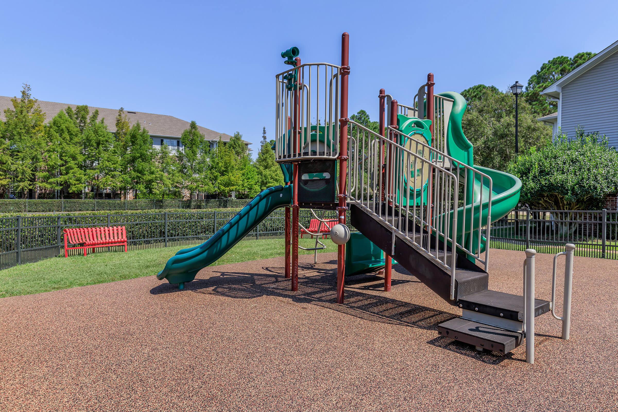 Playground equipment featuring a green slide, steps, and climbing structures, surrounded by grass and a red bench in the background. The scene is set in a well-maintained outdoor area with trees and a clear blue sky.