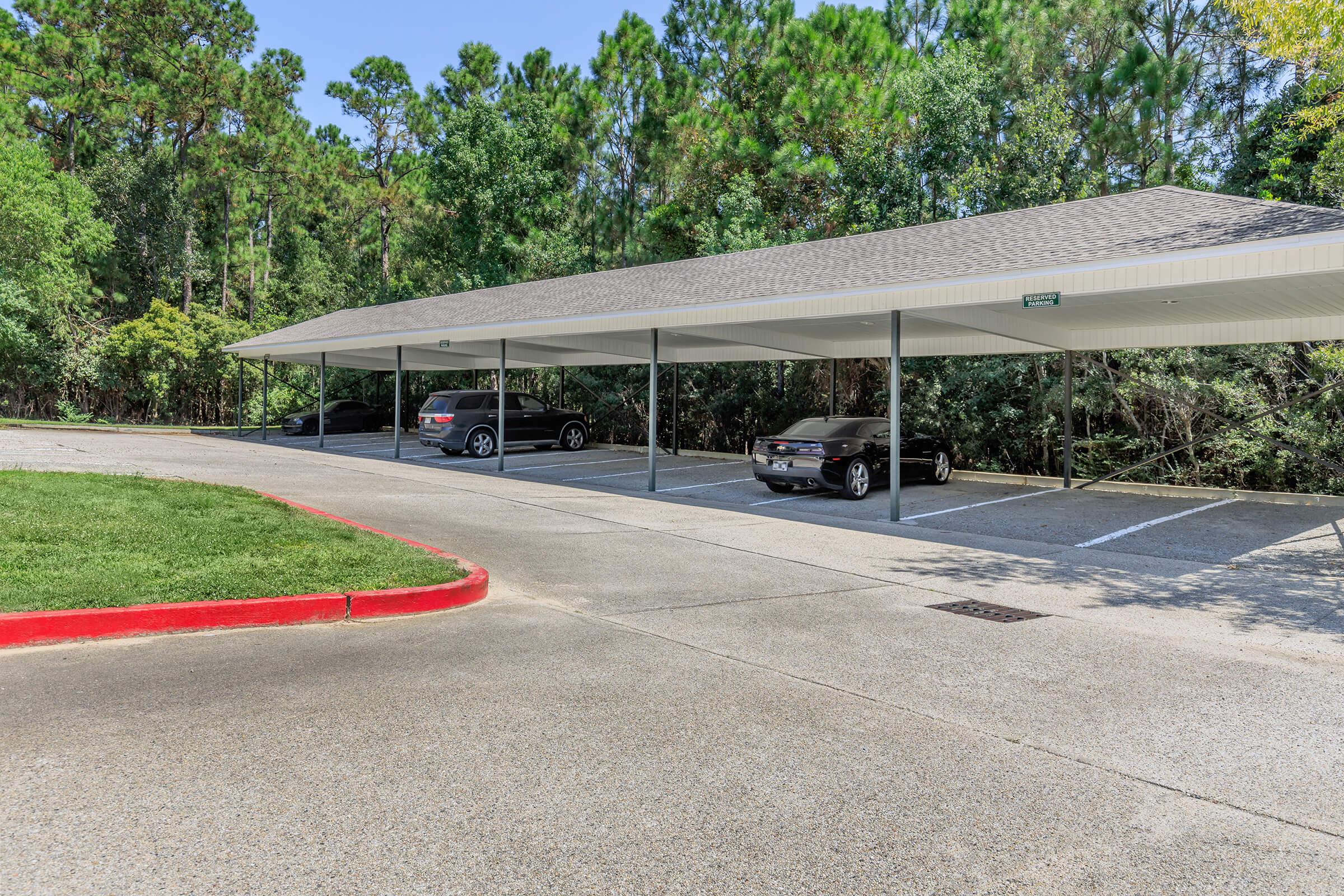 A covered parking area with two parked cars, surrounded by lush greenery and trees. The parking lot features a paved surface and a red curb along one side, leading to a path that curves gently. The structure has a sloped roof, providing shelter for the vehicles parked underneath.