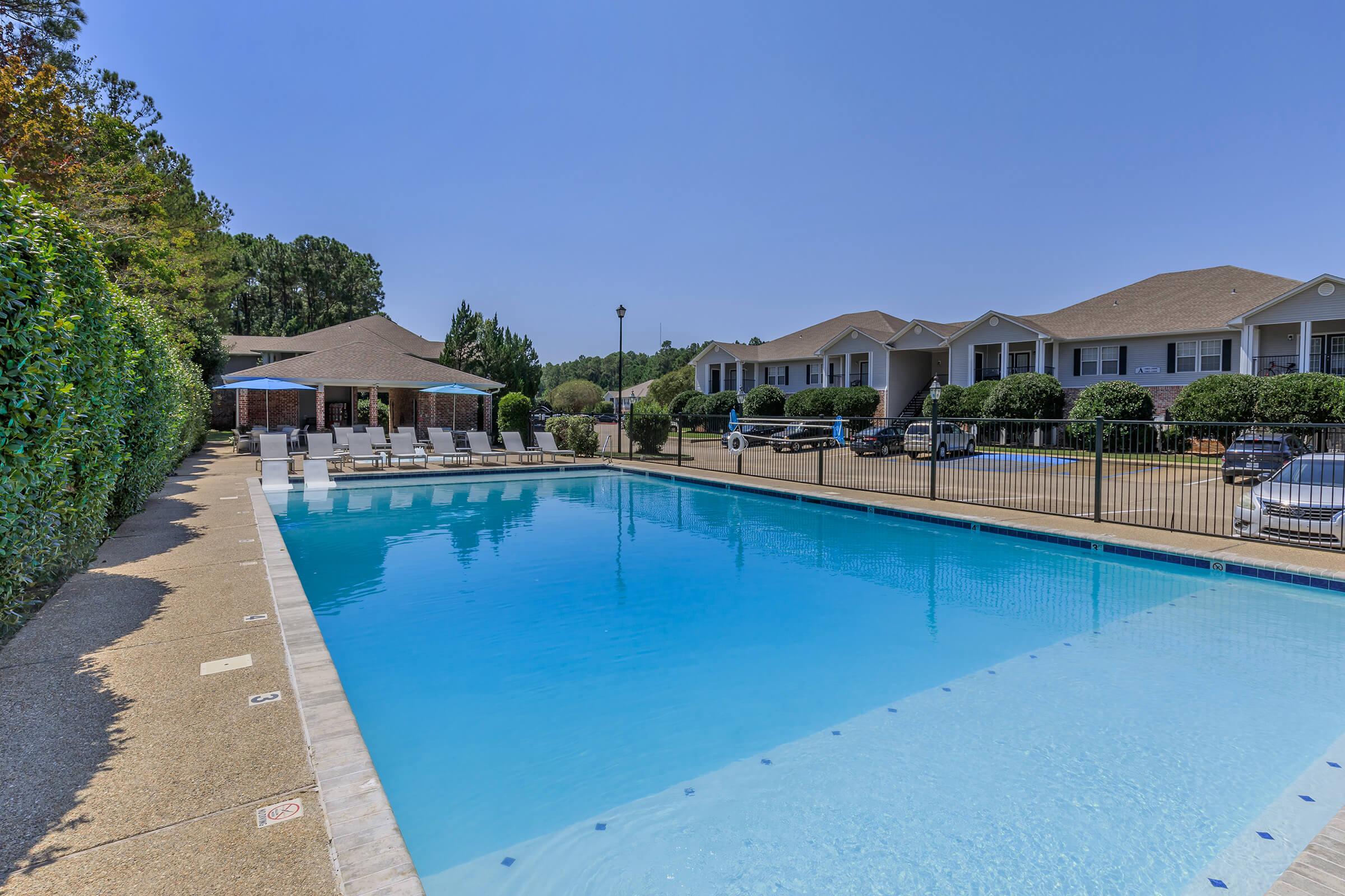 A clear blue swimming pool surrounded by lounge chairs and a grassy area. In the background, a pavilion with a seating area and several apartment buildings are visible under a sunny sky. A parking lot with cars is also visible nearby, framed by neatly trimmed hedges.