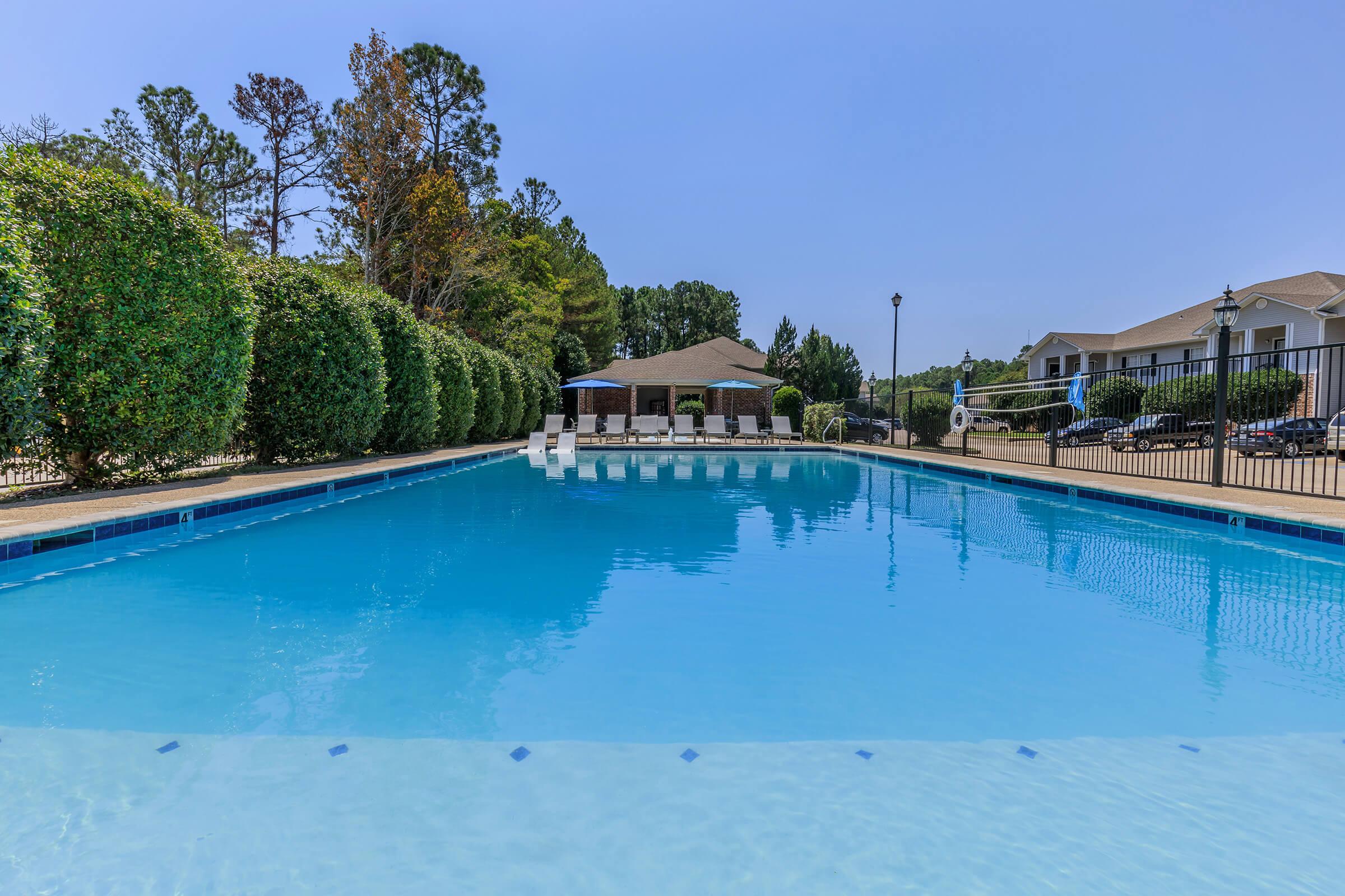 A serene swimming pool surrounded by neatly trimmed hedges and a clear blue sky. Sun loungers are positioned by the pool's edge, and a covered gazebo is visible in the background. The scene conveys a peaceful, inviting atmosphere perfect for relaxation.