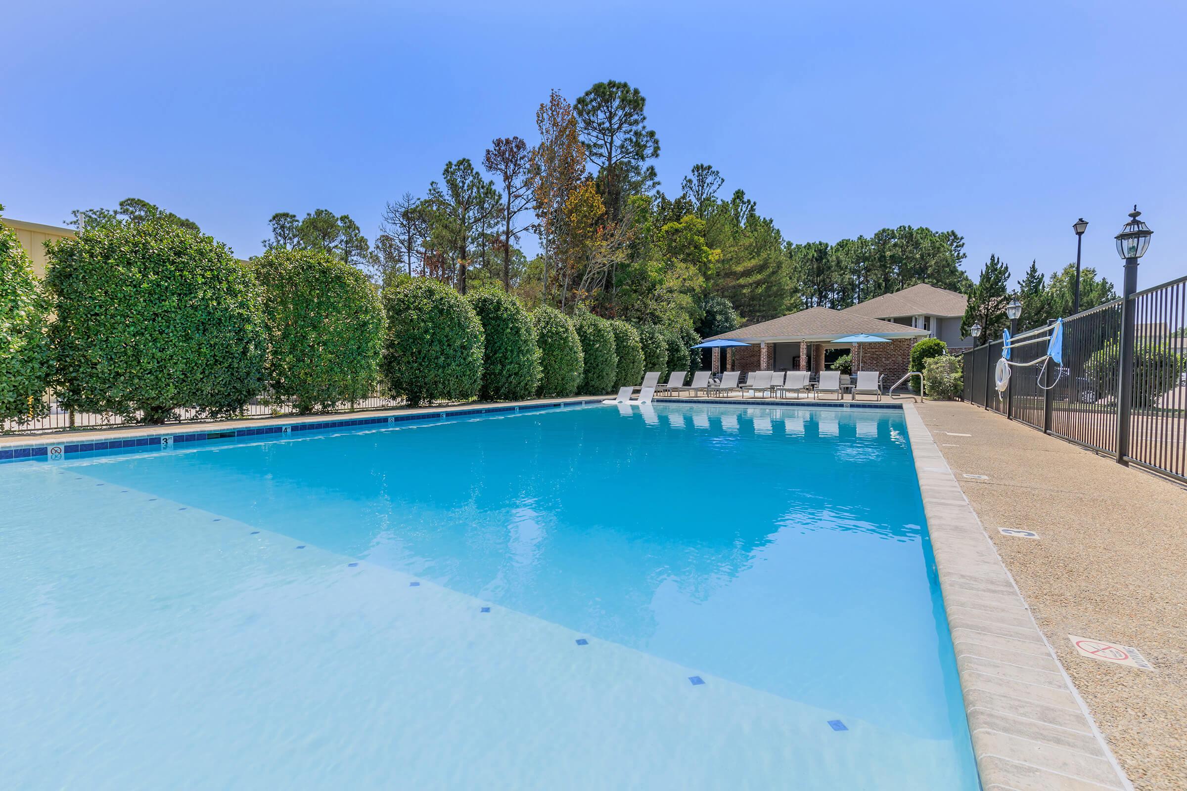A clear blue swimming pool surrounded by well-maintained hedges and lounge chairs. In the background, a covered pavilion and trees provide a serene atmosphere. The pool area is enclosed by a fence, and the sky is bright and clear. Perfect for relaxation and leisure activities.