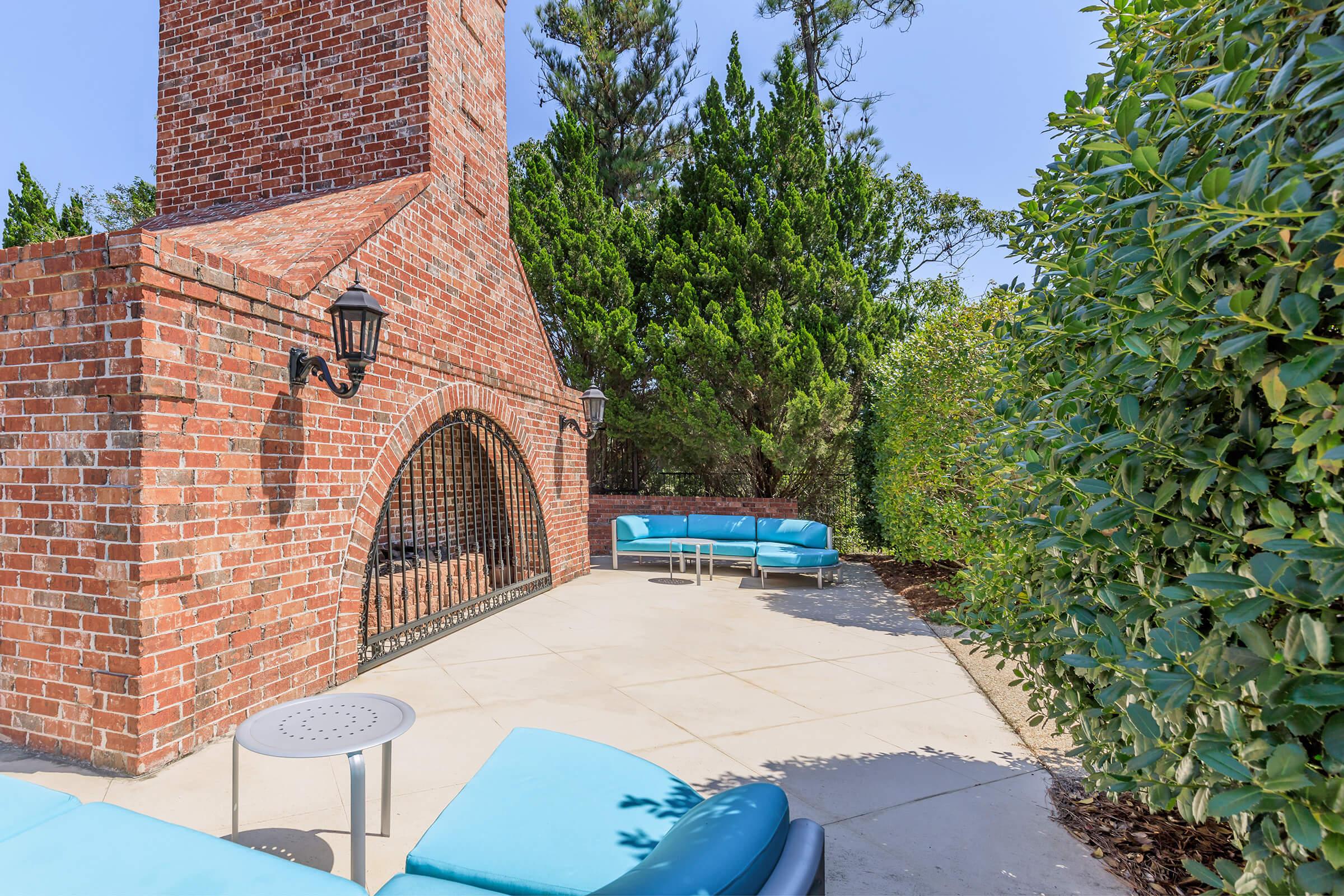 A patio area featuring a brick chimney and a small outdoor fireplace. There's a light blue seating arrangement with cushions and a circular table nearby. Lush greenery surrounds the space, providing a serene atmosphere. The sky is clear and sunny, enhancing the inviting outdoor setting.