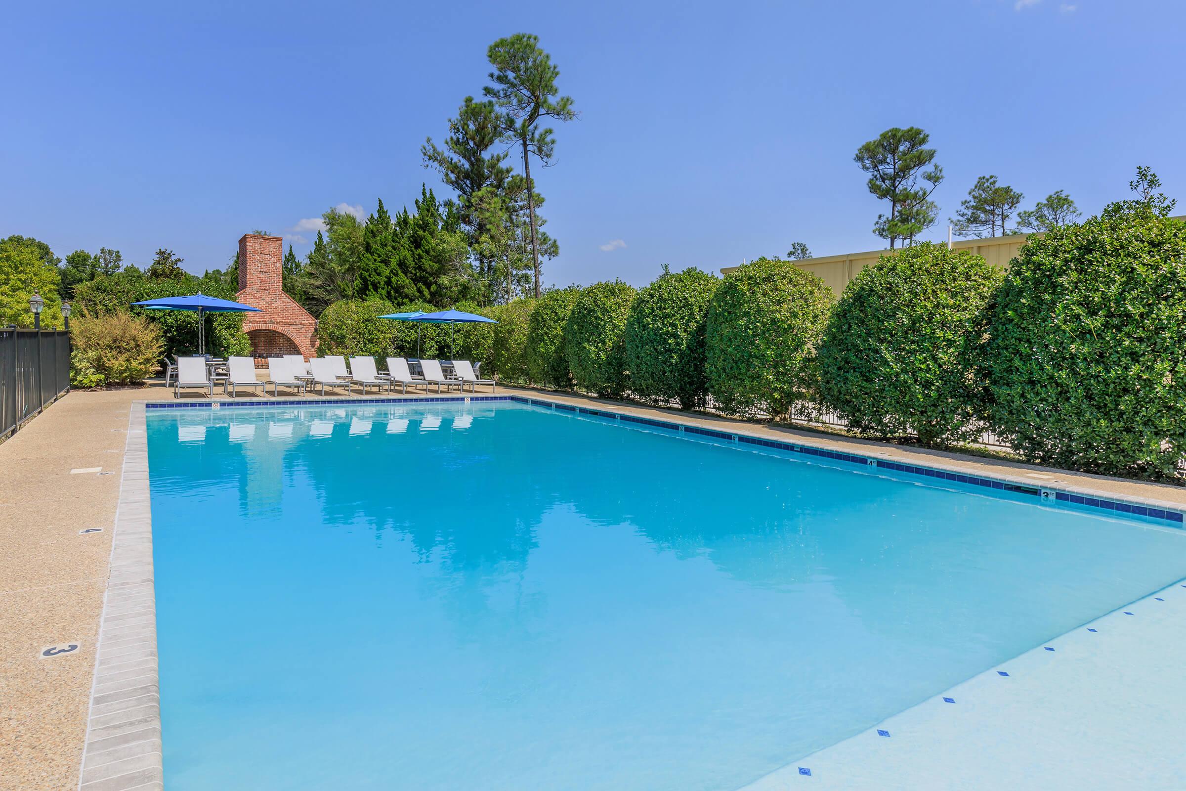 A clear blue swimming pool surrounded by lounge chairs and umbrellas, with lush green hedges lining the perimeter. In the background, a brick structure and tall trees are visible under a bright blue sky.