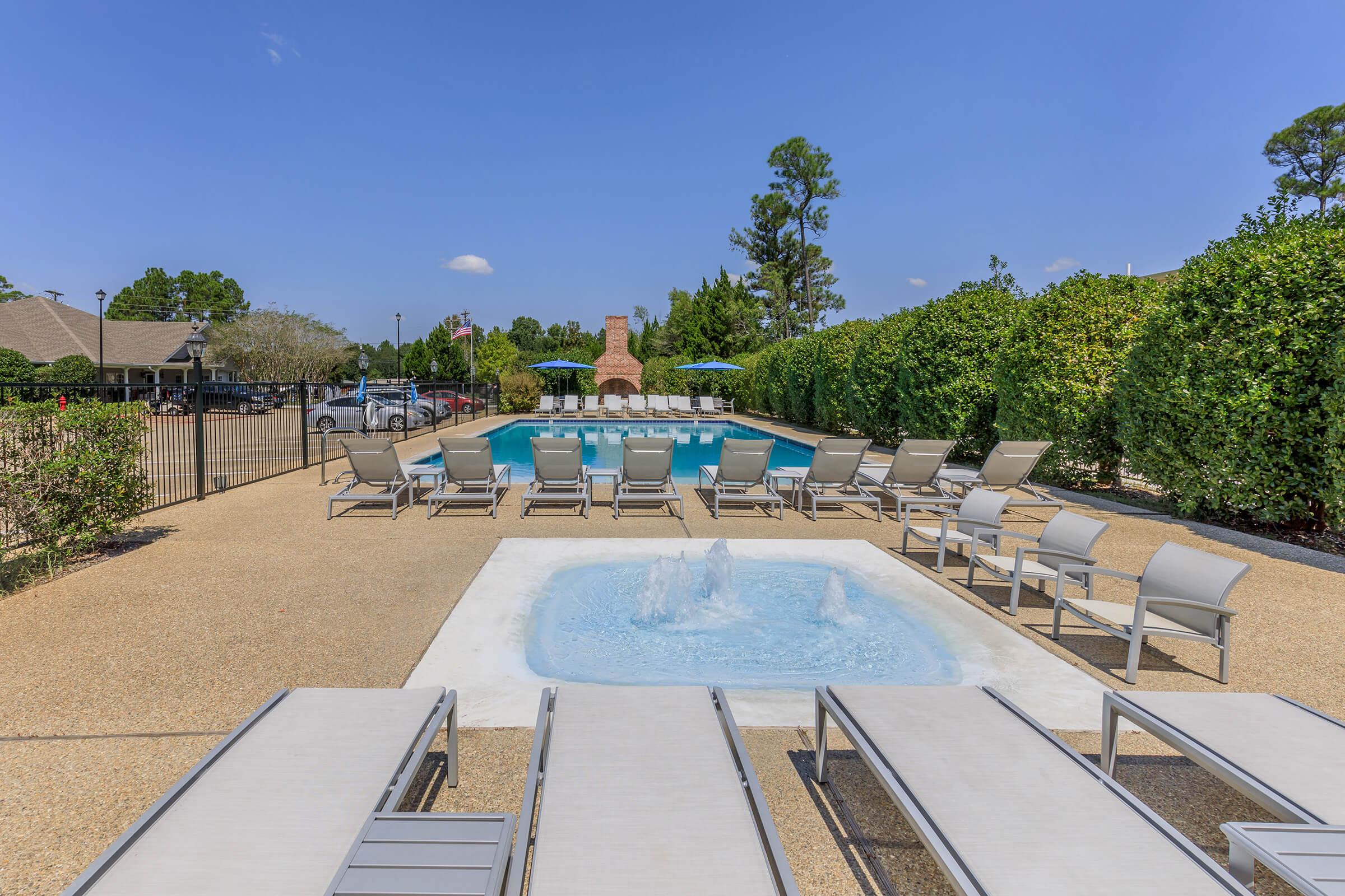A sunny outdoor pool area featuring a clear blue pool, surrounded by lounge chairs and patio umbrellas. In the foreground, a small fountain adds decor to the space, while a well-maintained hedge provides privacy. The setting is ideal for relaxation and leisure activities.