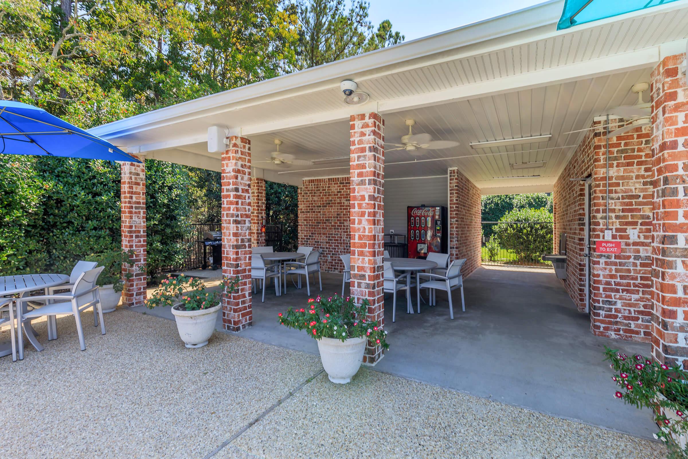 Covers outdoor seating area with brick columns, ceiling fans, and tables. A blue umbrella provides shade. Potted flowers adorn the space, and a vending machine is visible nearby. Lush greenery surrounds the area, creating a relaxing atmosphere.