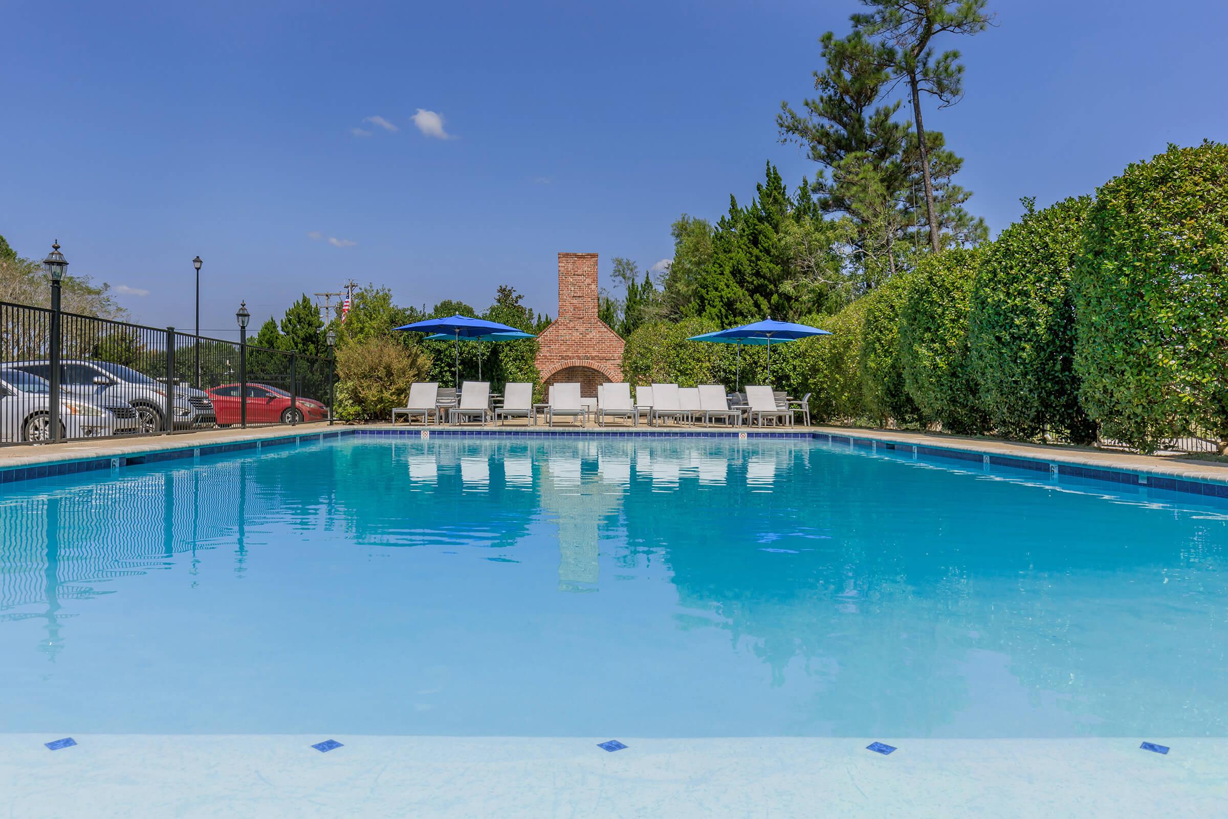 A clear blue swimming pool surrounded by lounge chairs and umbrellas, with a brick fireplace in the background. Green trees and shrubs line the pool area, and a few cars are parked nearby, under a bright blue sky with a few clouds.