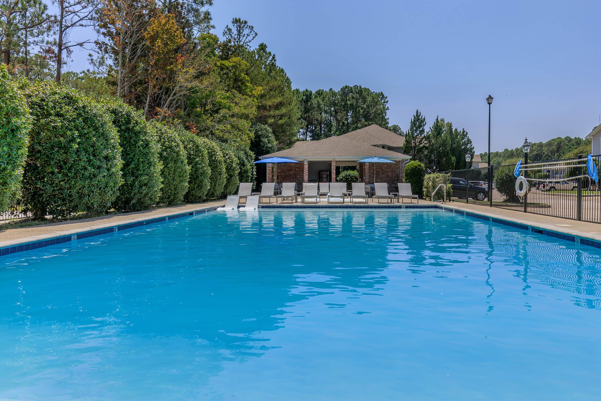 A clear blue swimming pool surrounded by neatly trimmed hedges and trees. In the background, there is a shaded area with lounge chairs and umbrellas. The scene is set on a sunny day with a clear sky, creating a serene and inviting atmosphere for relaxation.