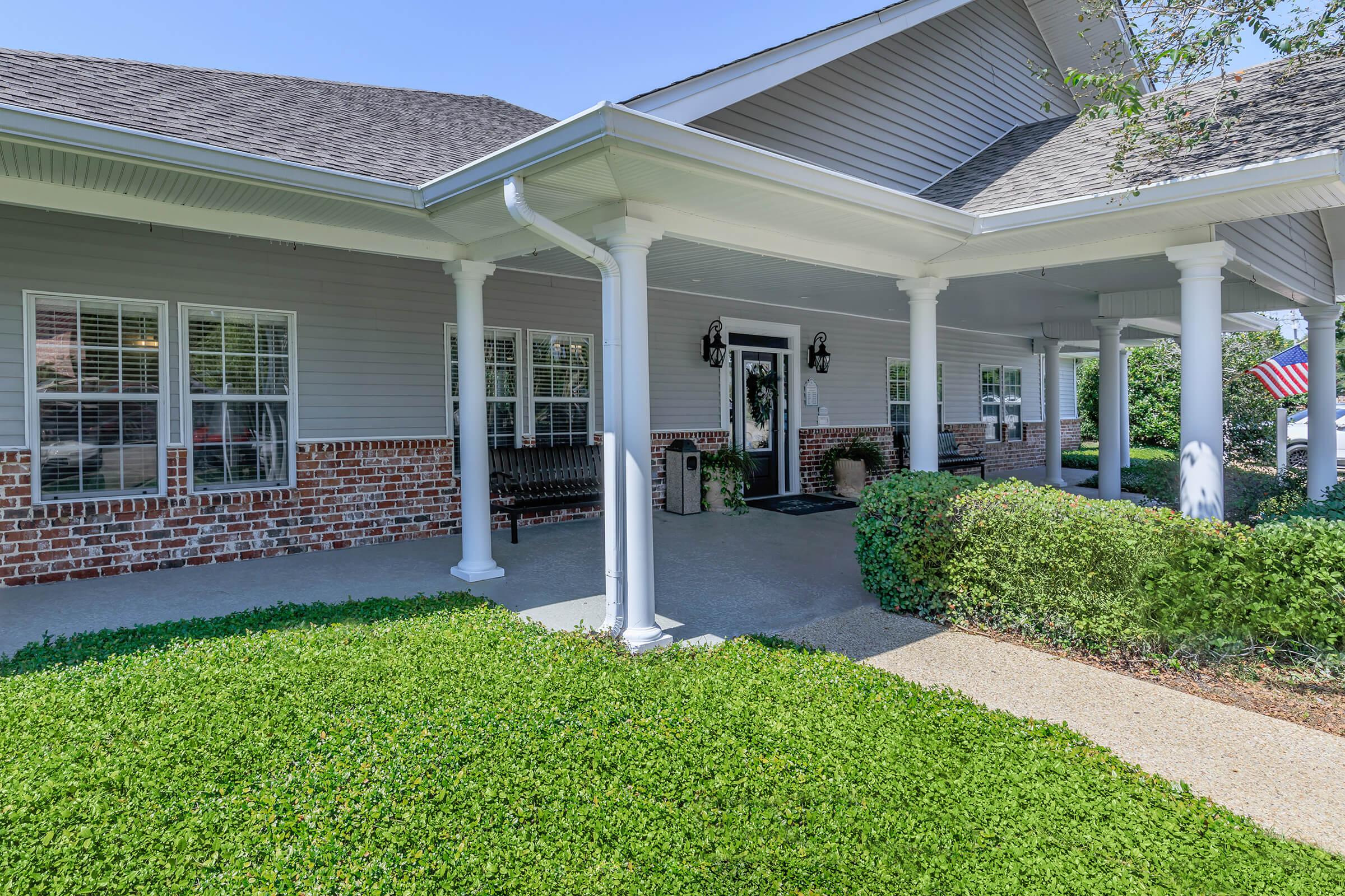 A welcoming front porch of a residential building featuring white columns, benches, and a neatly trimmed green hedge. The facade is a mix of light gray siding and brick, with several windows allowing natural light. An American flag is visible in the background, adding a patriotic touch to the scene.