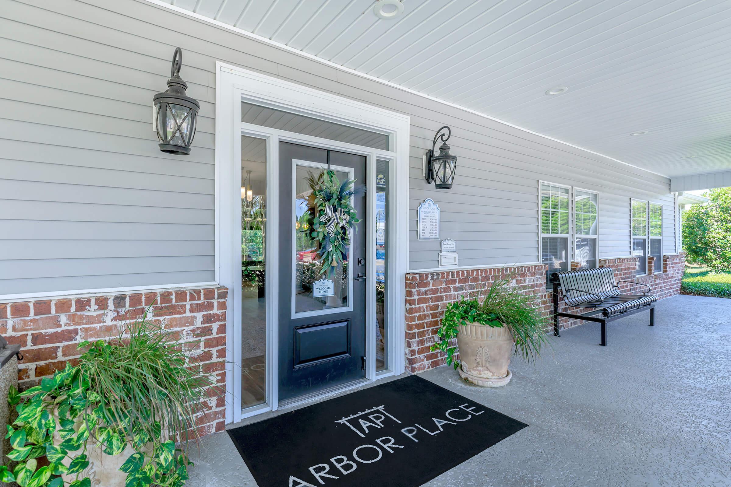 Entrance of a building named "Arbor Place" with a decorative wreath on the door. The facade features gray siding, brick accents, and large windows. There’s a bench on the porch and potted plants, creating a welcoming atmosphere. Two lantern-style lights flank the door under a covered area.
