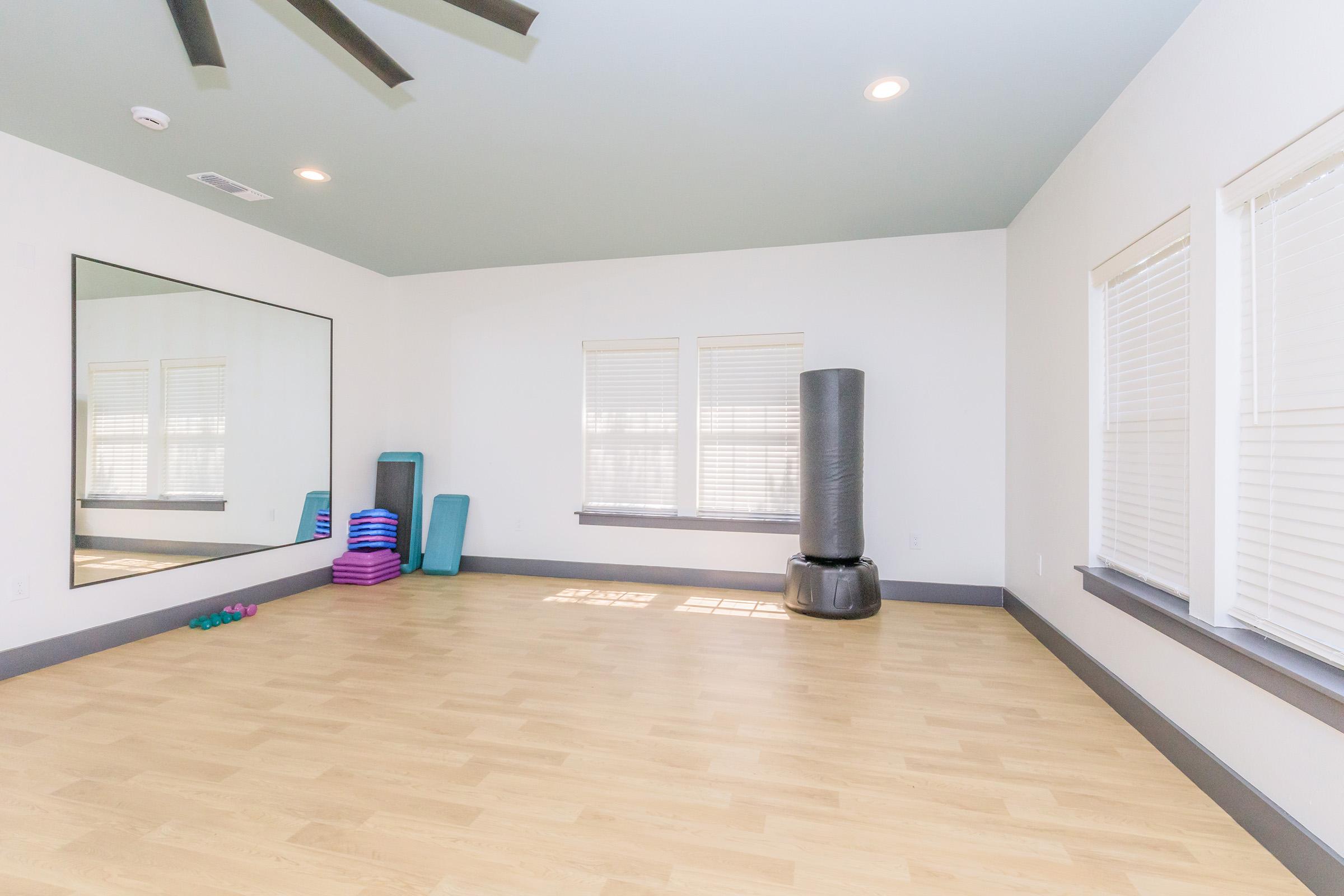 A spacious, empty exercise room featuring a large mirror, a black punching bag, colorful exercise mats stacked, and small weights on the wooden floor. Natural light comes through multiple windows with blinds, and the ceiling has a fan. The walls and ceiling are painted in light, neutral tones.
